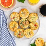 A plate of fluffy vegetable egg muffins, crafted from a classic egg muffin cups recipe, is surrounded by a glass of orange juice, a cup of coffee, and a halved grapefruit. A blue striped napkin is also visible on the side. The muffins are golden-brown with visible diced vegetables.