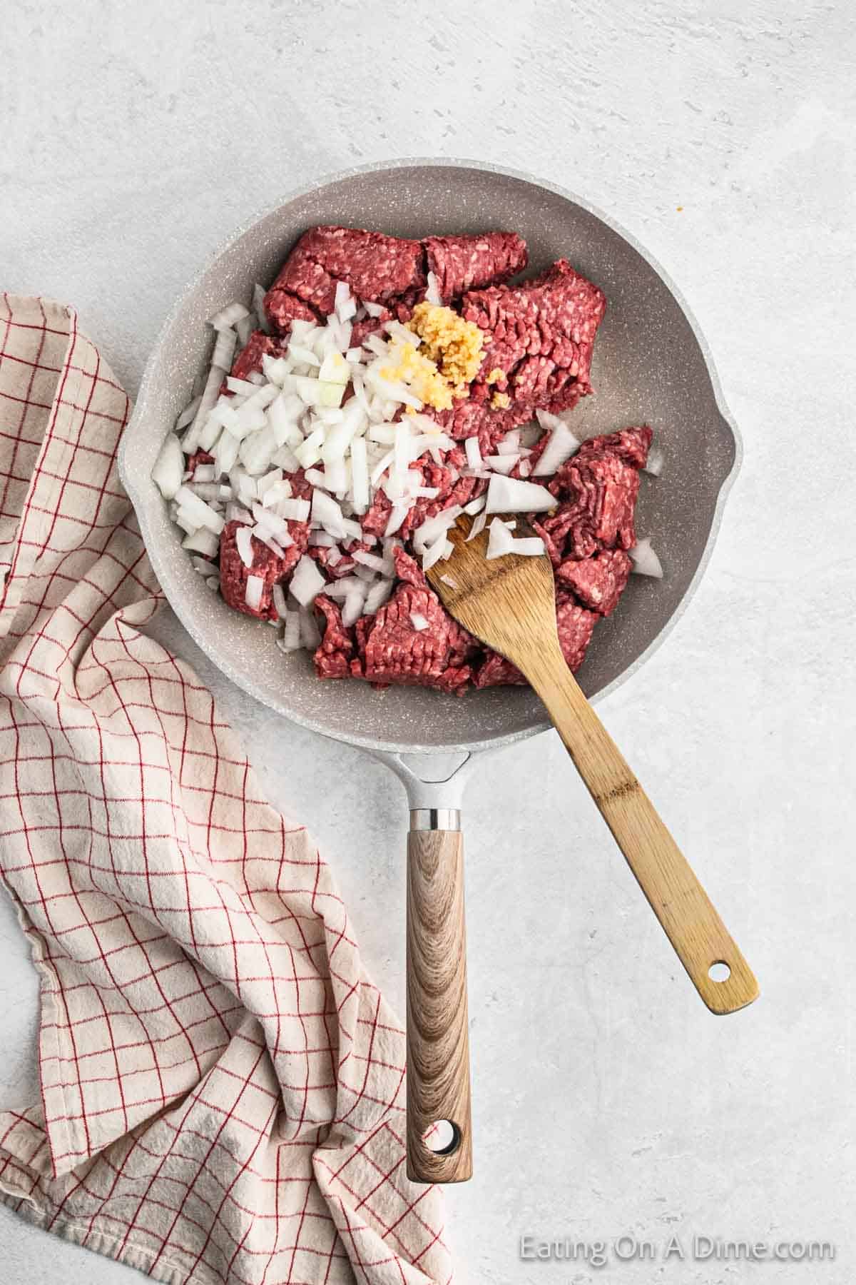 Cooking ground beef in a large skillet with chopped onions and minced garlic with a wooden spoon