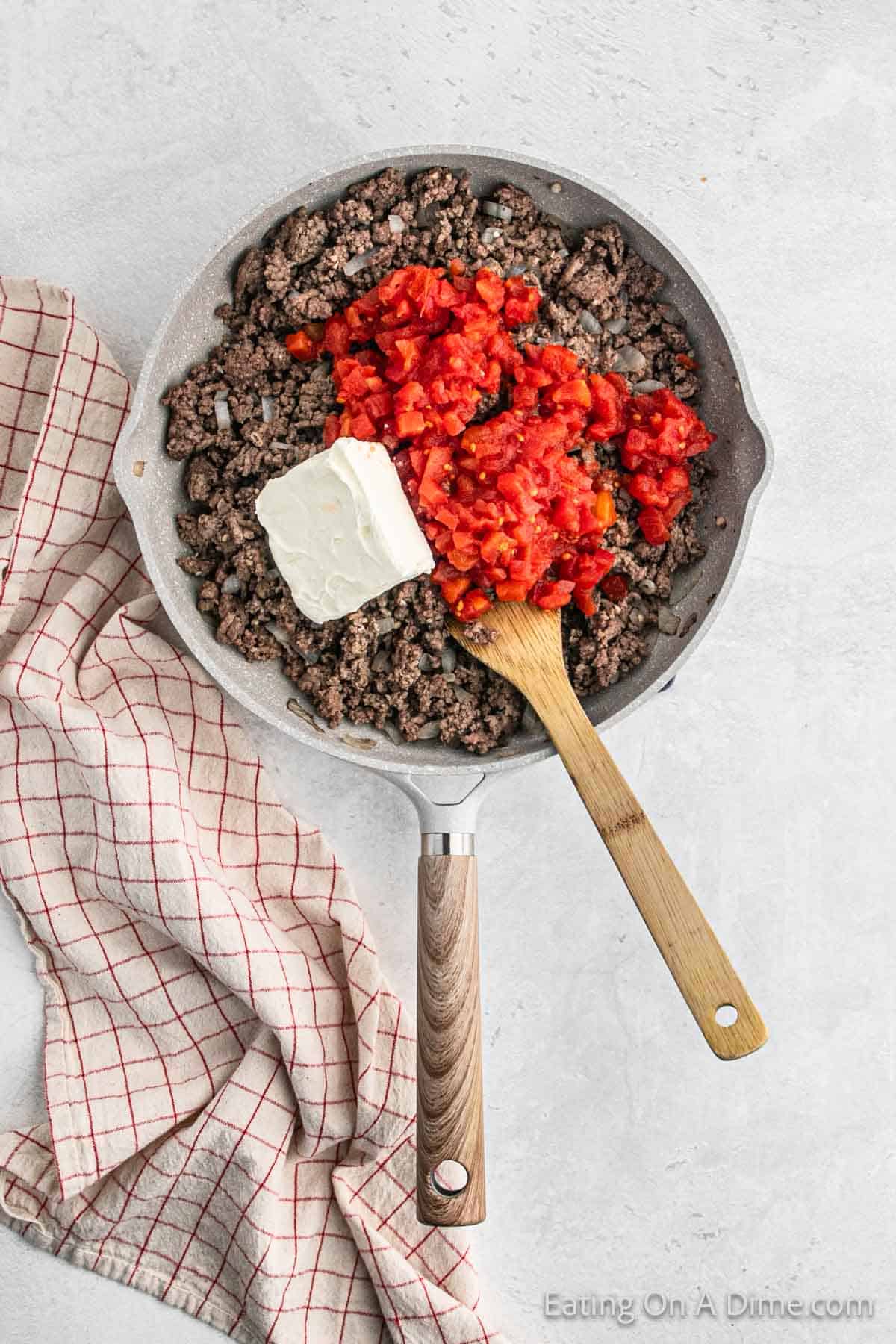 Cooked ground beef in a skillet with diced tomatoes and cream cheese with a wooden spoon