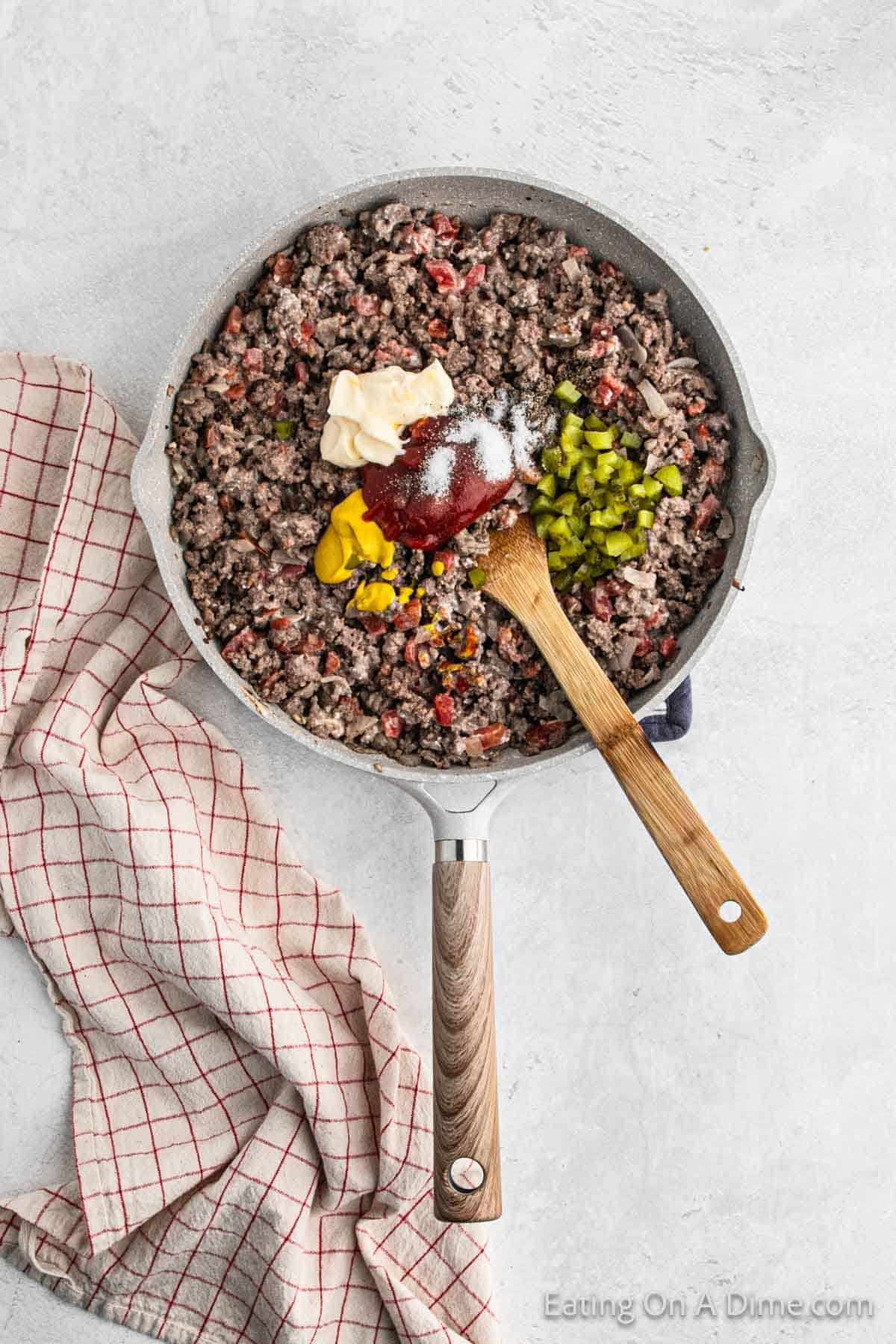 Cooked ground beef in a large skillet with mayonnaise, ketchup, diced pickles, mustard and seasoning with a wooden spoon