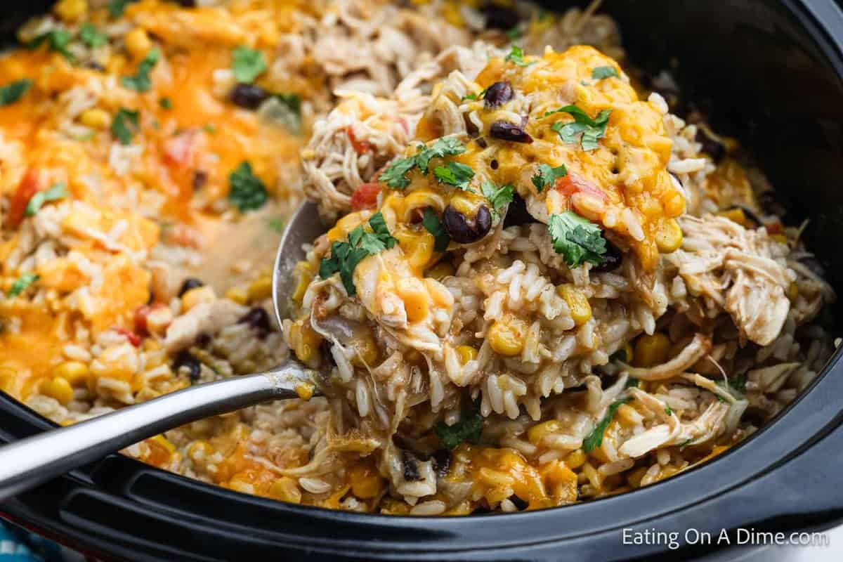 A close-up of a spoonful of cheesy Southwest crock pot chicken and rice loaded with black beans, corn, and herbs, being lifted from a slow cooker filled with the dish. The background shows more of the flavorful, cheesy Mexican-inspired meal. Text at the bottom reads, "Eating On a Dime.com".