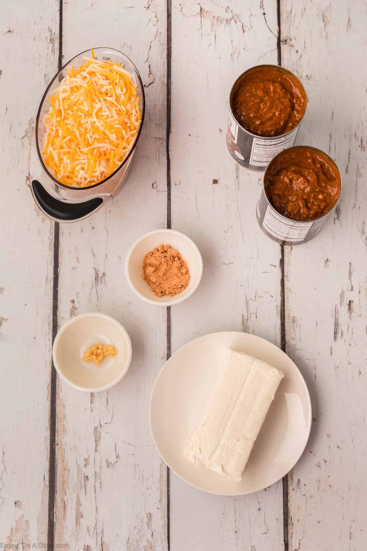 A flat lay of ingredients for a chili cheese dip recipe on a wooden surface. Includes a bowl of shredded cheese, two opened cans of chili, a white dish with a block of cream cheese, a small bowl of brown sugar, and a small bowl of minced garlic.