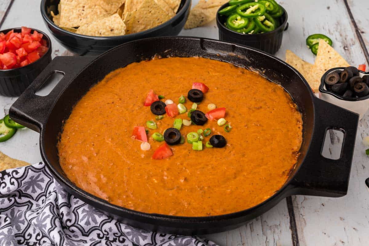 A skillet filled with a creamy, orange-colored chili cheese dip recipe topped with diced tomatoes, sliced black olives, and chopped green onions. Surrounding the skillet are bowls of tortilla chips, jalapeño slices, diced tomatoes, and sliced black olives. A patterned napkin is beside the skillet.