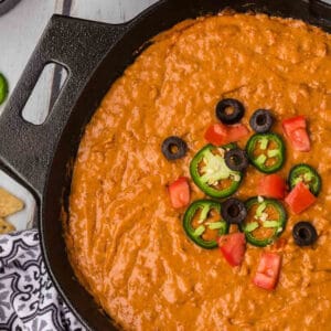 A cast iron skillet filled with creamy refried beans topped with sliced jalapeños, black olives, and diced tomatoes, reminiscent of a favorite chili cheese dip recipe. The skillet is placed on a wooden surface with a patterned cloth partially visible in the corner.