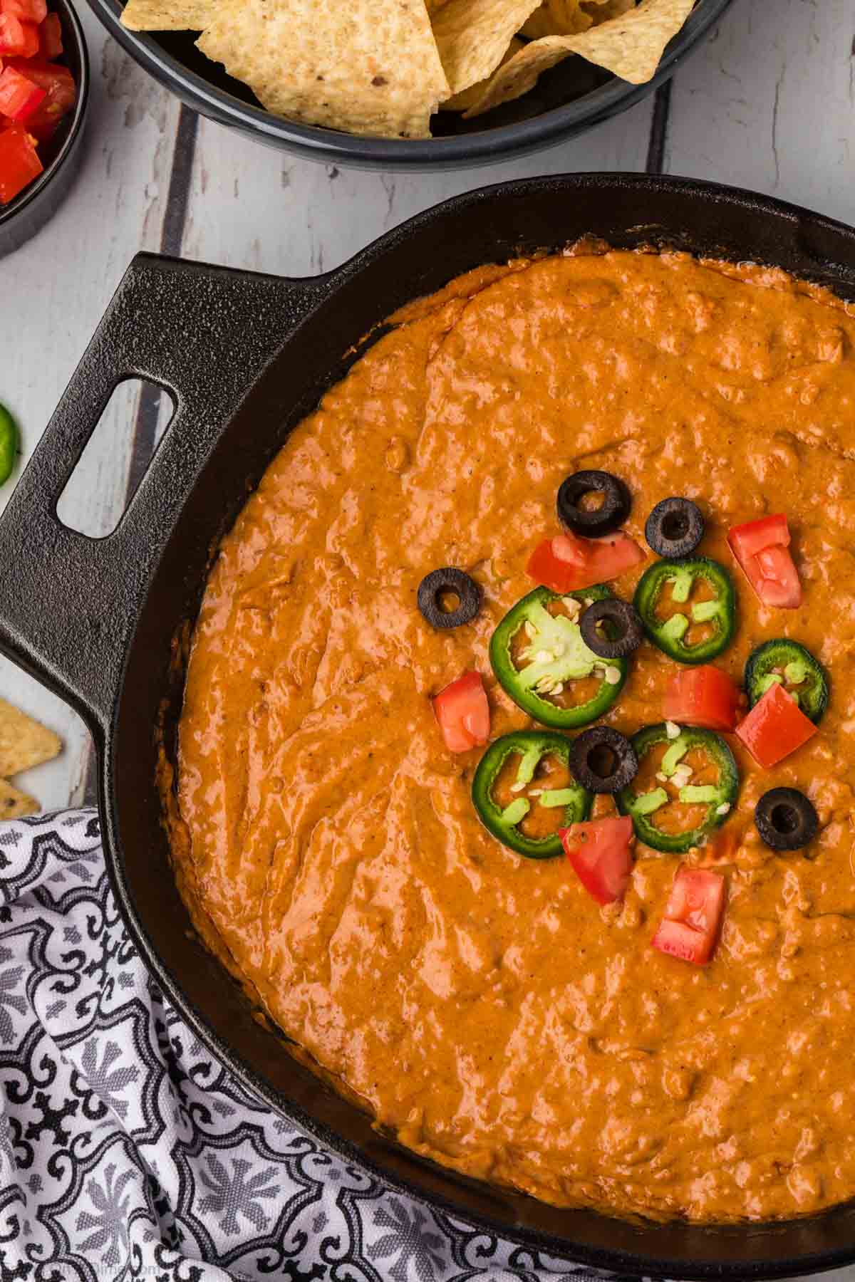 A skillet filled with creamy, orange-hued chili cheese dip, garnished with sliced jalapeños, black olives, and diced tomatoes. A dish with tortilla chips and a small bowl of diced tomatoes are placed nearby, while a patterned napkin is situated on the bottom left corner.