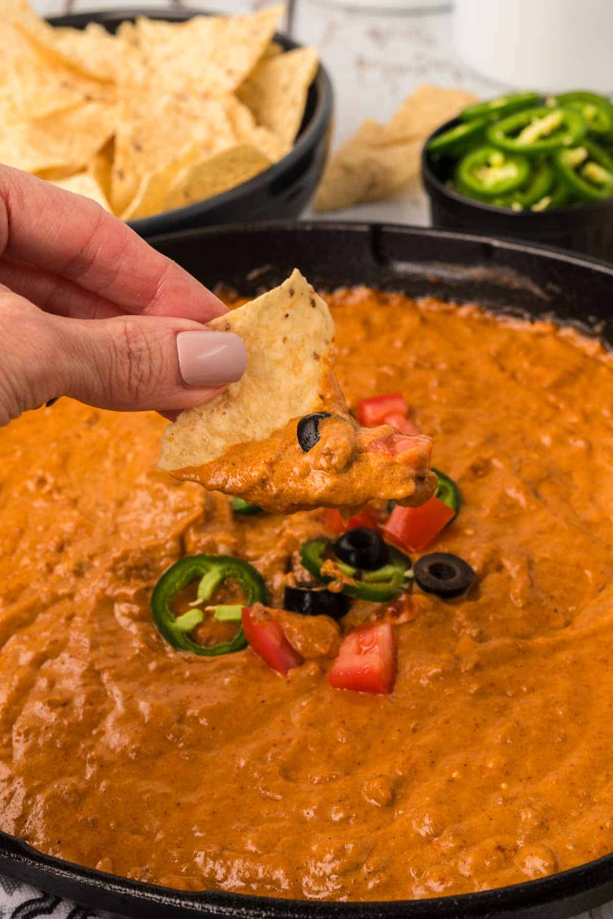 A hand dips a tortilla chip into a chili cheese dip, garnished with slices of jalapeño, diced tomatoes, and black olives, in a black dish. A bowl of tortilla chips and a bowl of sliced jalapeños are visible in the background.