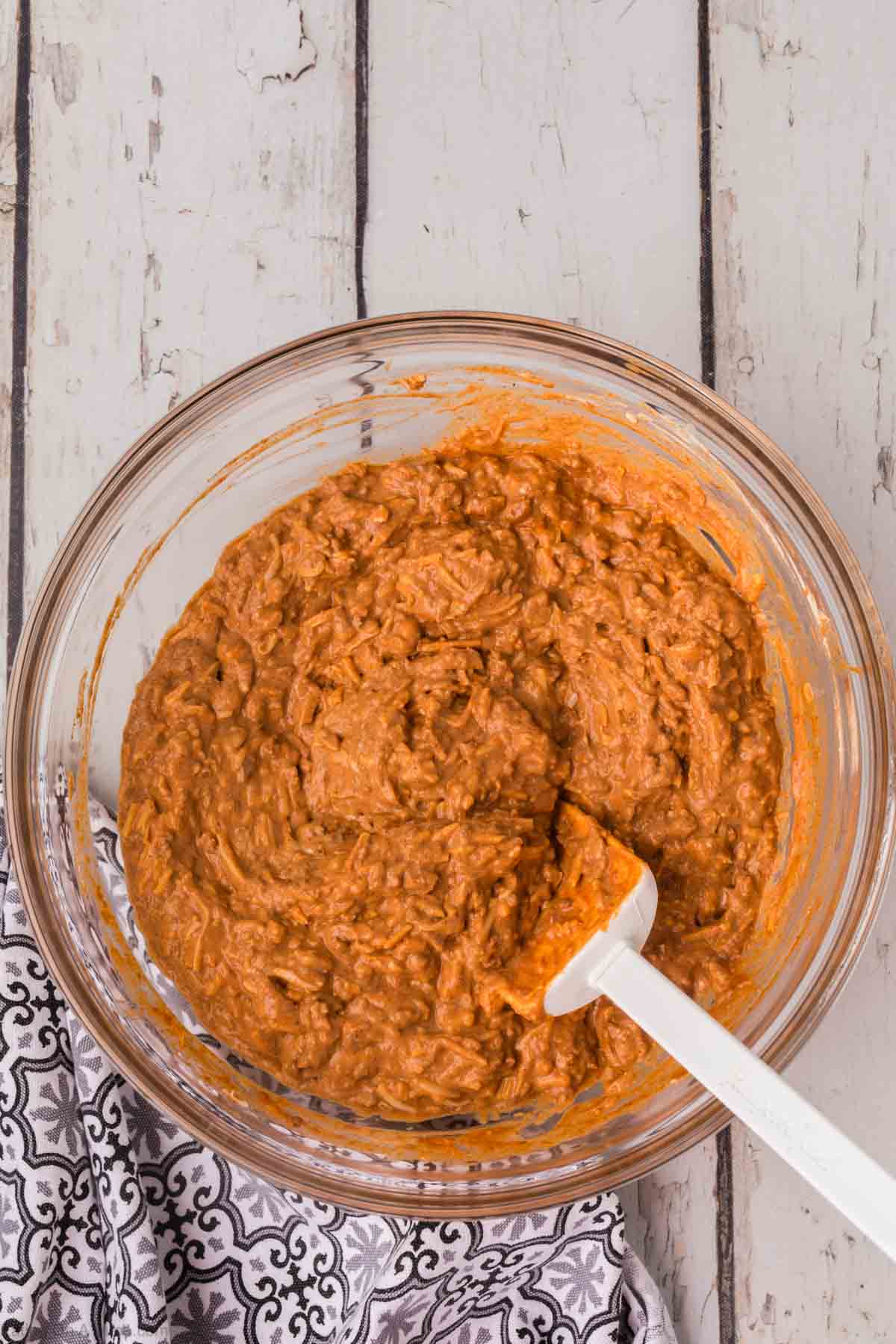 A glass mixing bowl filled with thick, orange hummus mixture, resembling a chili cheese dip recipe, placed on a distressed wooden surface. A white spatula is resting inside the bowl. A portion of a patterned black and white cloth napkin is visible on the bottom left.