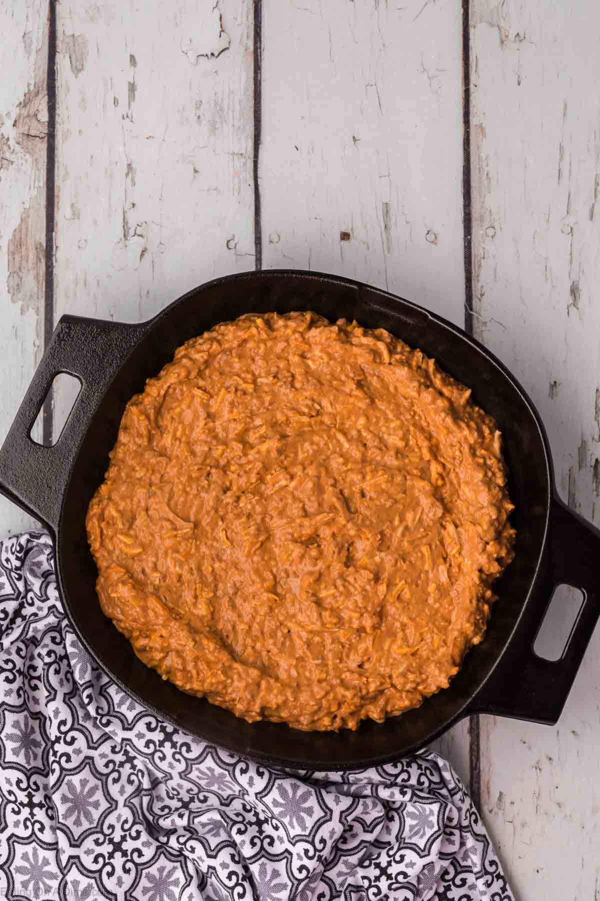 A cast iron skillet filled with a thick, orange-colored chili cheese dip sits on a wooden surface. The dish appears creamy and textured. Part of a patterned black and white cloth is visible next to the skillet.