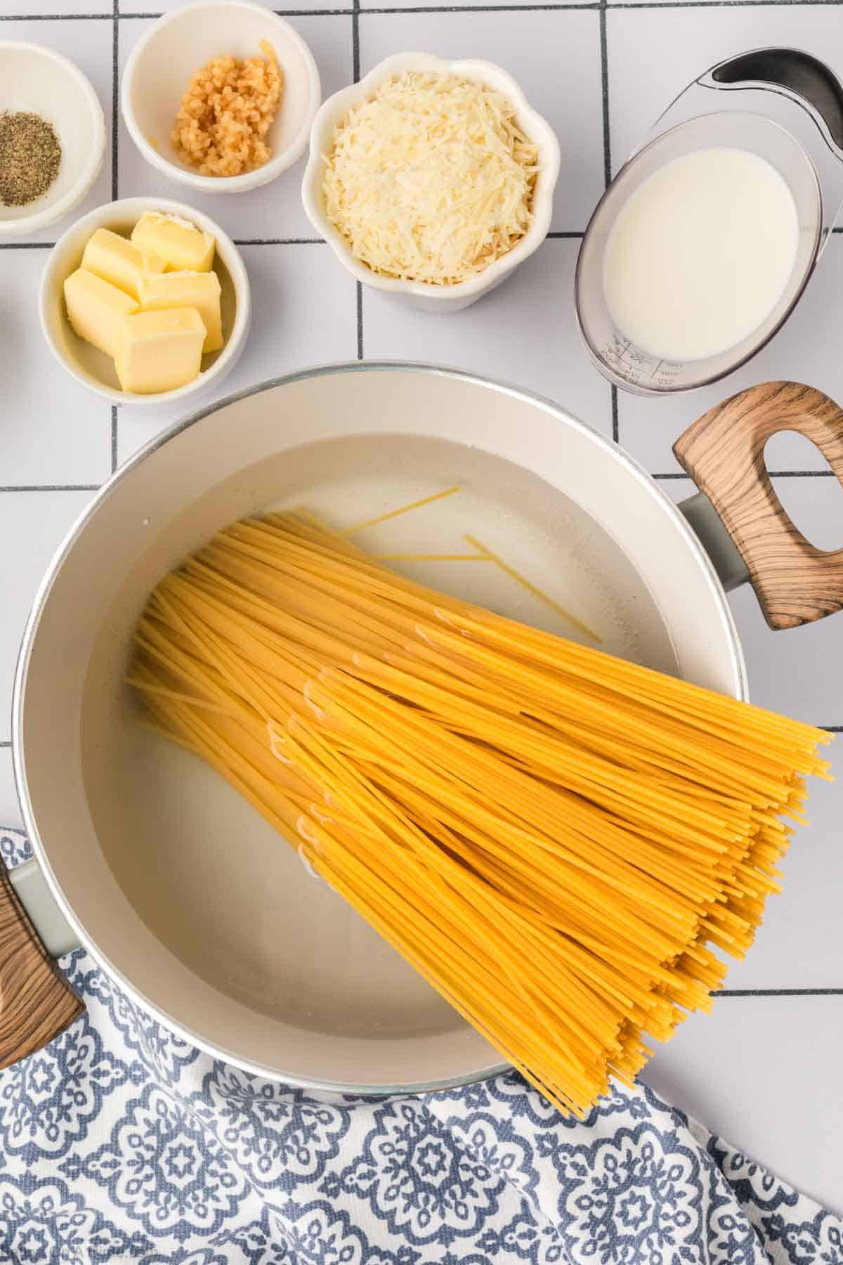 Spaghetti Noodles in a large pot of water with small bowls of butter, parmesan cheese, garlic and pepper on the side