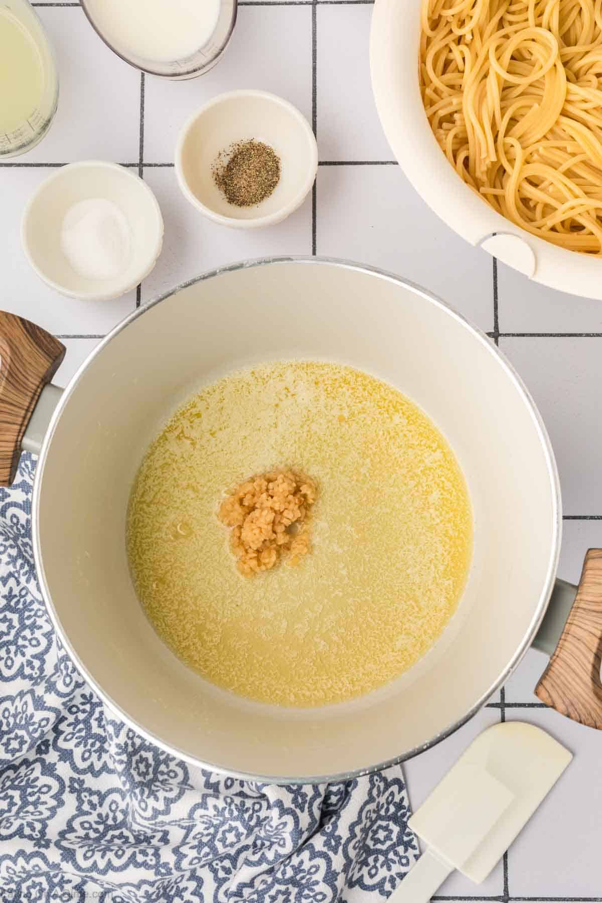 Melted butter in a large pot with minced garlic with a bowl of pasta and seasoning on the side