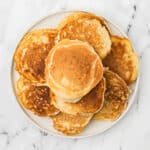 A plate of golden brown pancakes stacked neatly on a white marble surface exudes warmth. With a fluffy texture and varying shades, they appear freshly cooked. Curious about preserving them? Explore how to freeze pancakes to savor their goodness anytime.