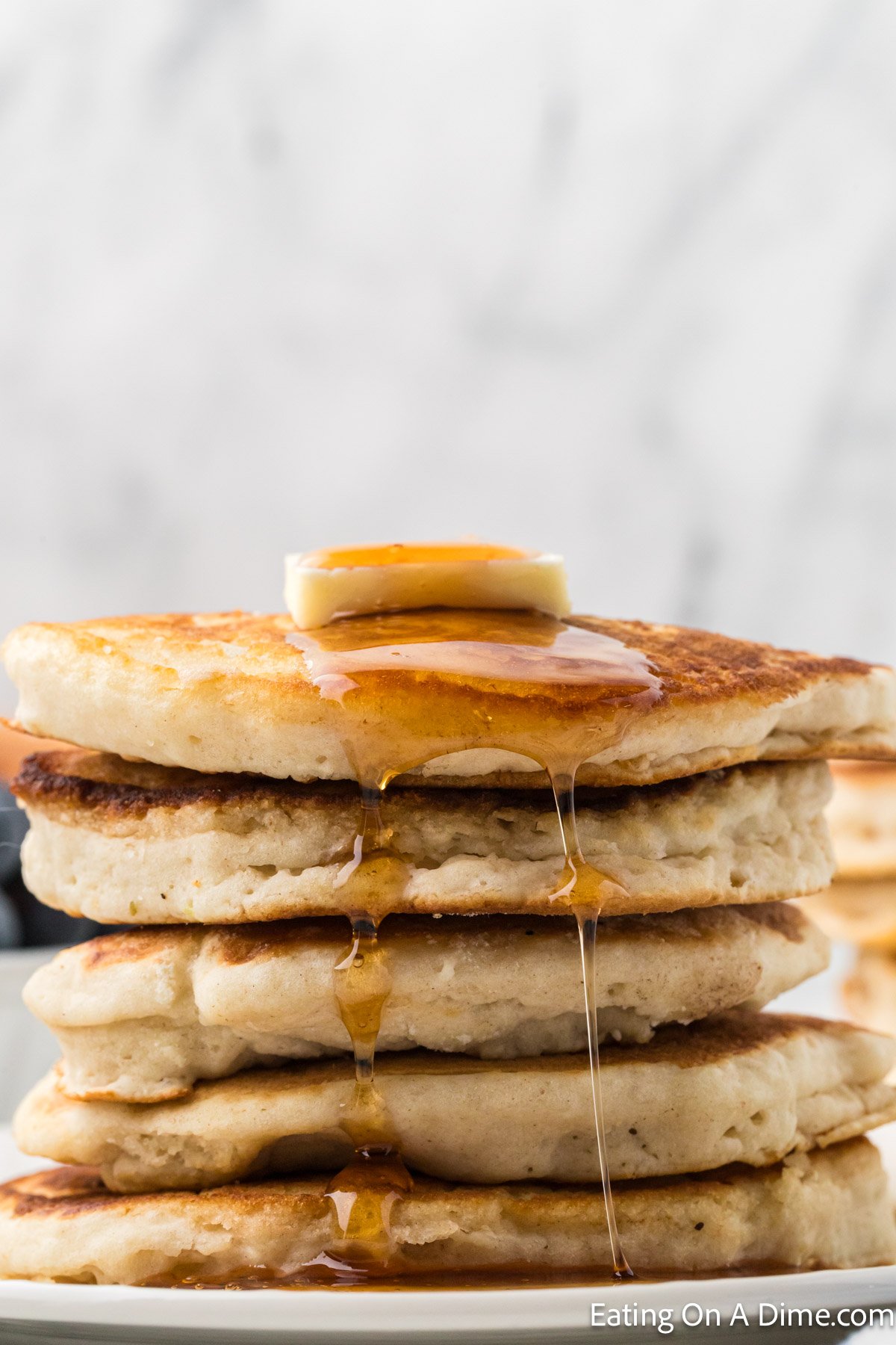 A stack of fluffy pancakes, perfect for brunch or learning how to freeze pancakes later, is topped with a pat of butter and has golden syrup dripping down the sides. The background features a soft, blurred marble texture.