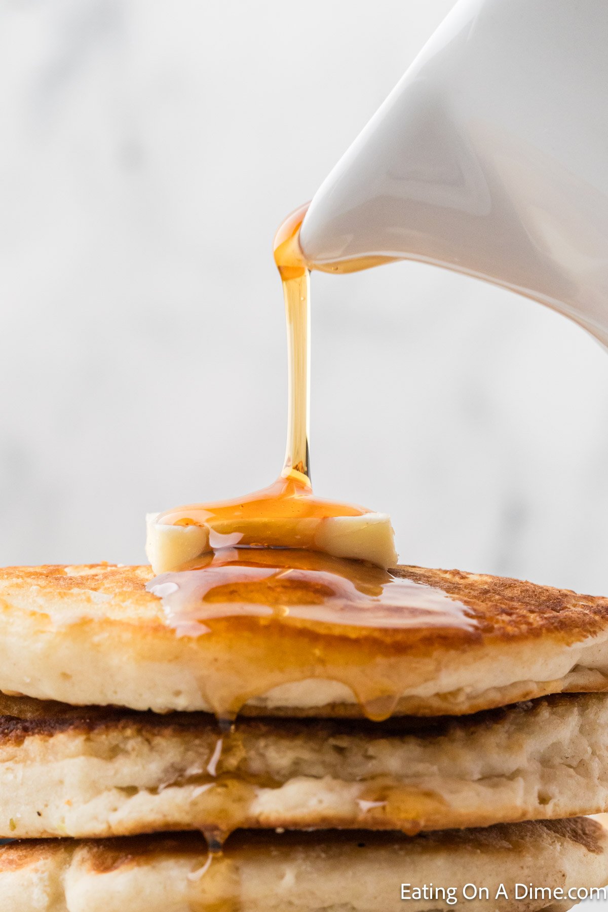 A stack of fluffy pancakes, perfect for learning how to freeze pancakes later, is topped with a pat of butter and drizzled with syrup from a white pitcher. The syrup cascades down the sides, creating a mouthwatering breakfast scene against a light background.