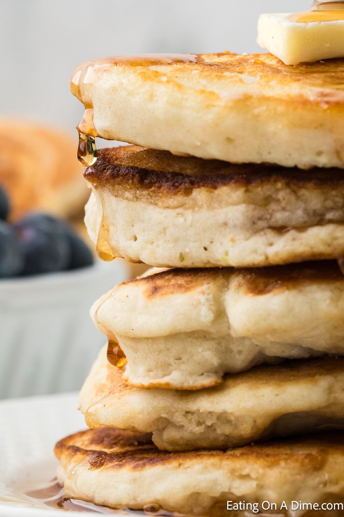 A close-up of a stack of fluffy pancakes topped with a pat of butter, syrup cascading down the sides. A bowl of blueberries is blurred in the background—a snapshot before finding out how to freeze pancakes for another delicious morning.