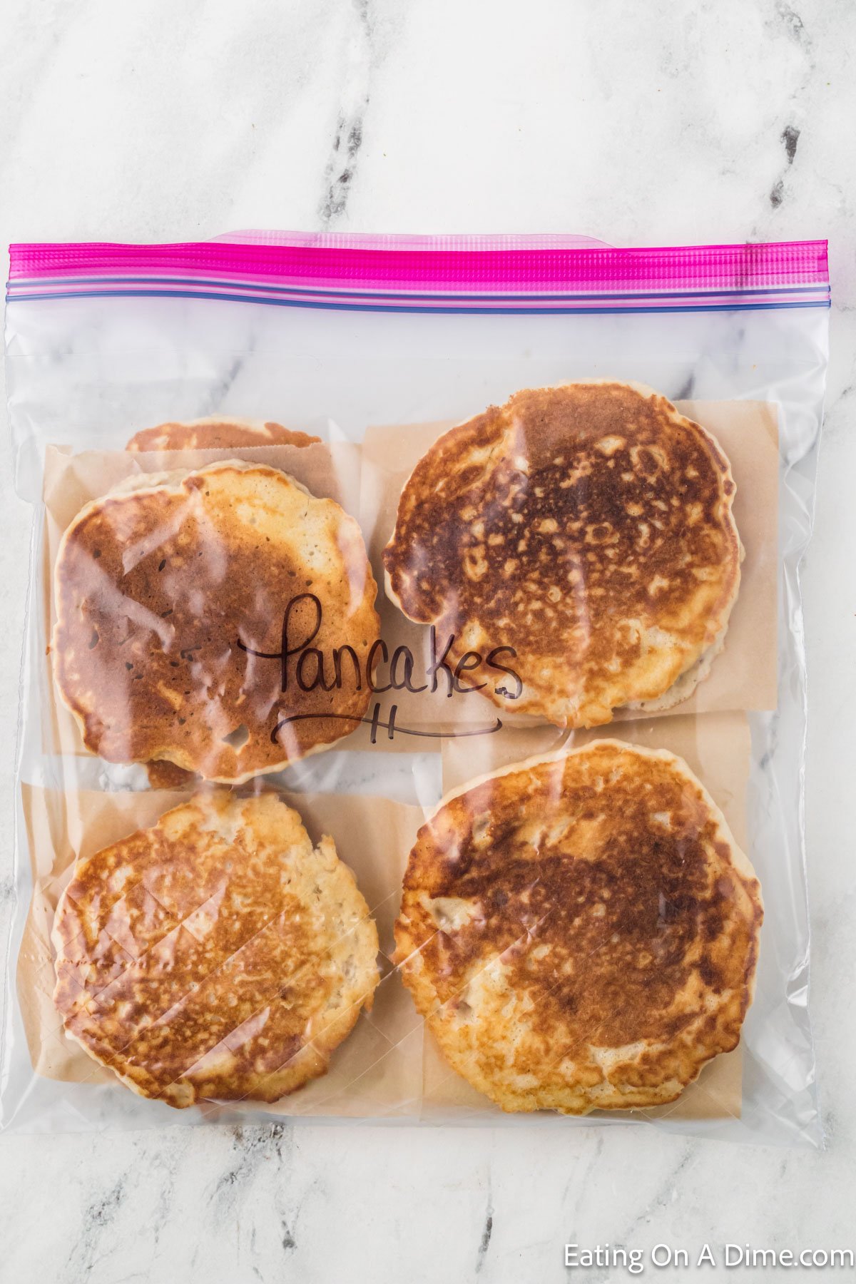 Four pancakes stored in a resealable plastic bag labeled "Pancakes" lie on a marble surface, ready to teach you how to freeze pancakes effectively. Each pancake is separated by parchment paper, and the bag is sealed with a pink zipper seal, ensuring freshness.