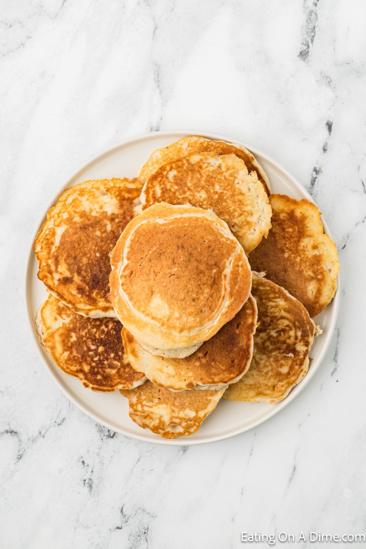 A plate of golden-brown pancakes stacked on a marble countertop invites indulgence. The pancakes, perfect for any morning, vary slightly in size and boast a fluffy texture. Wondering how to freeze pancakes? Simply stack them with parchment in between, ensuring deliciousness any day of the week.