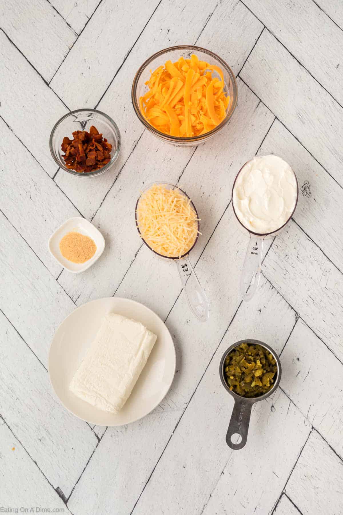 Ingredients for a delicious Jalapeño Popper Dip Recipe arranged on a white wooden surface: a block of cream cheese on a plate, a bowl of shredded cheddar cheese, cooked bacon bits, grated parmesan, sour cream, chopped jalapeños, and a small dish of garlic powder.