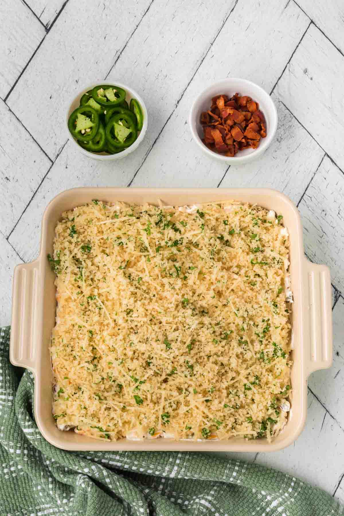 A dish filled with a casserole topped with breadcrumbs and herbs sits on a textured white surface, embodying the flavors of a Jalapeño Popper Dip. Nearby are small bowls of sliced jalapeños and diced bacon. A green checkered cloth is partially visible at the bottom.