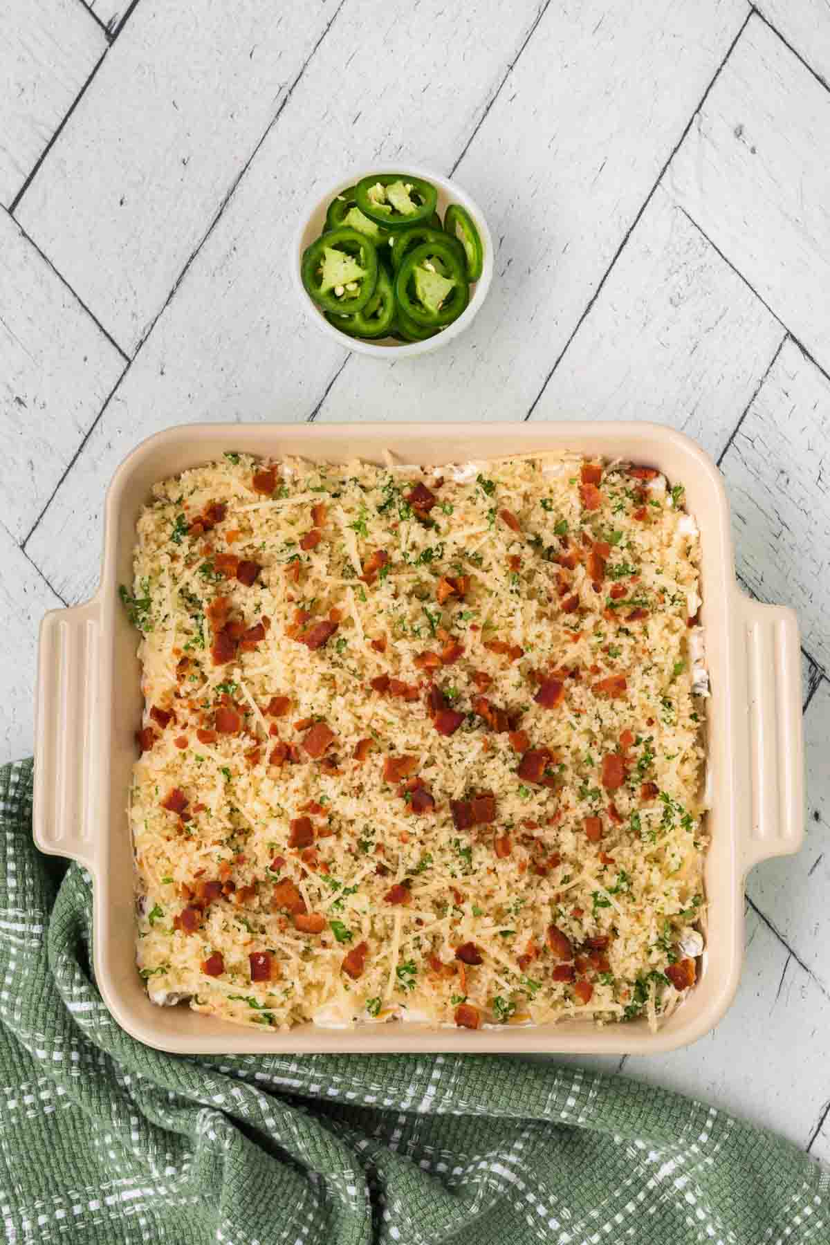 A baked casserole, reminiscent of a Jalapeño Popper Dip recipe, topped with breadcrumbs, cheese, herbs, and bacon bits in a rectangular dish on a white herringbone-patterned surface. A small bowl of sliced jalapeños is nearby, alongside a green checkered cloth.