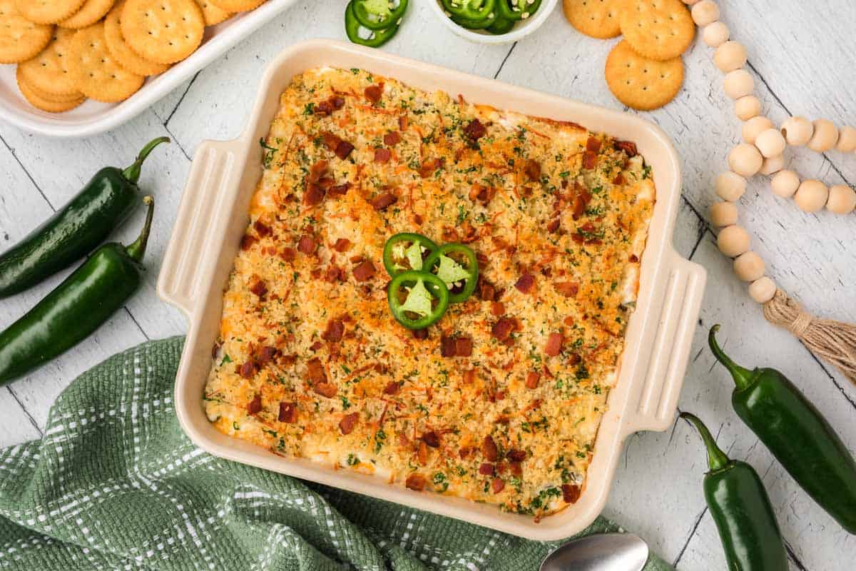 A square dish of jalapeño popper dip recipe topped with sliced jalapeños and bacon sits on a wooden table. Surrounded by whole jalapeños, round crackers, and a green checkered cloth.