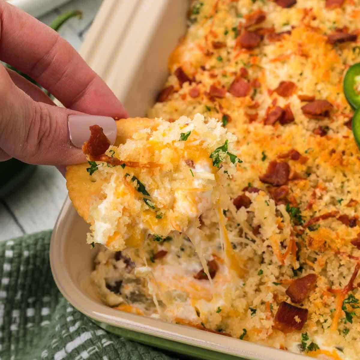 A person is holding a cracker dipped in a creamy Jalapeño Popper Dip Recipe. The dip, nestled in a baking dish, is topped with crispy breadcrumbs, melted cheese, bacon bits, and garnished with sliced jalapeños and herbs.