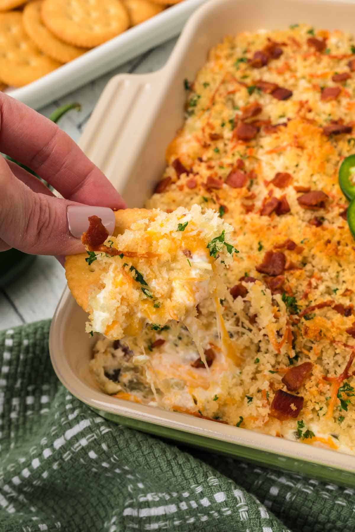 A hand dips a cracker into a creamy, cheesy Jalapeño Popper Dip, baked and topped with breadcrumbs, bacon bits, and herbs. This flavorful dish sits in a rectangular pan on a green cloth, with extra crackers visible in the background.