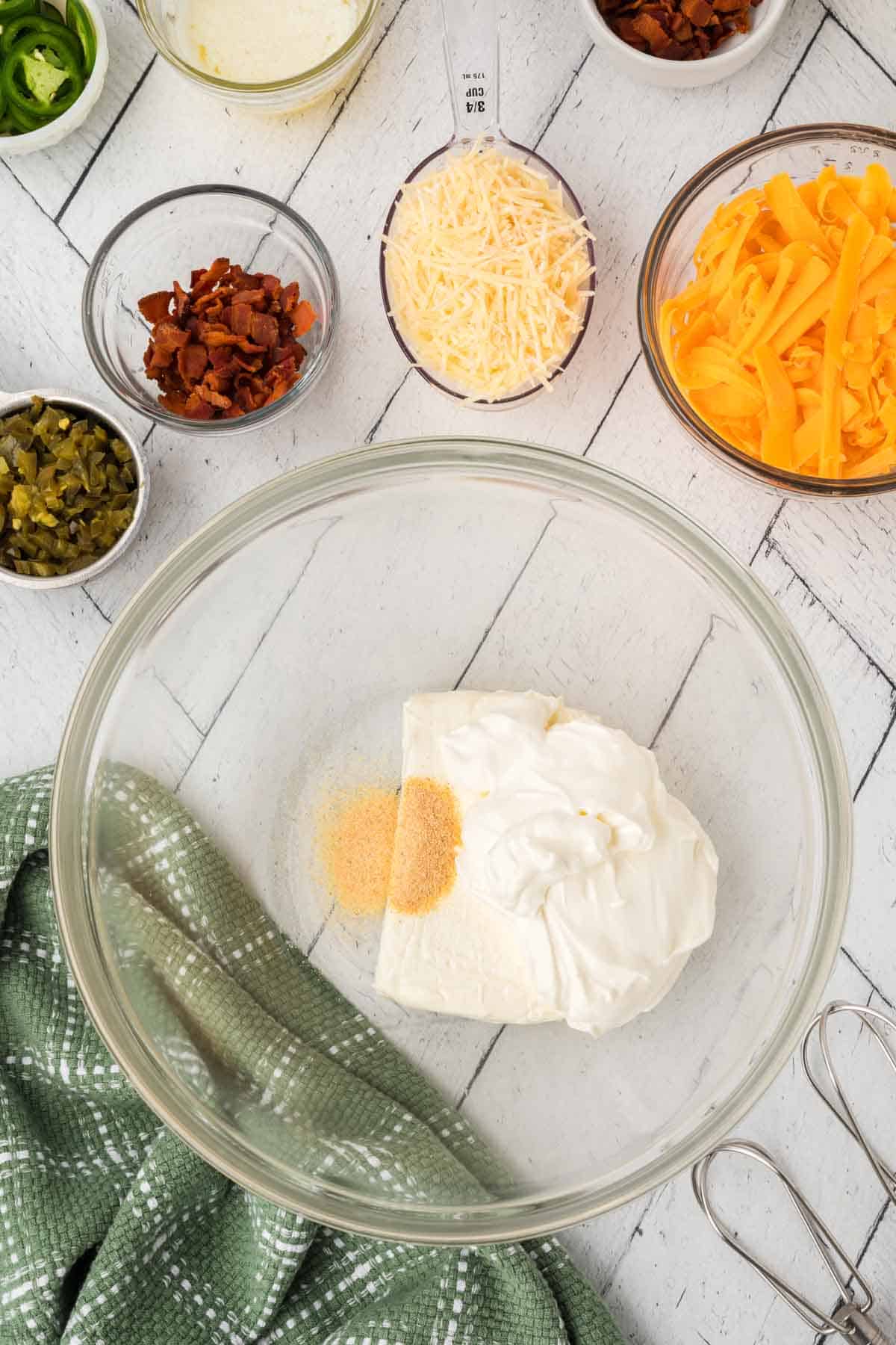 A clear bowl with cream cheese, sour cream, and seasoning sits on a white wooden surface, embodying the essence of a Jalapeño Popper Dip Recipe. Surrounding it are small bowls of bacon bits, shredded cheese, Parmesan, jalapeños, and pickles. A green kitchen towel and a metal whisk are nearby.