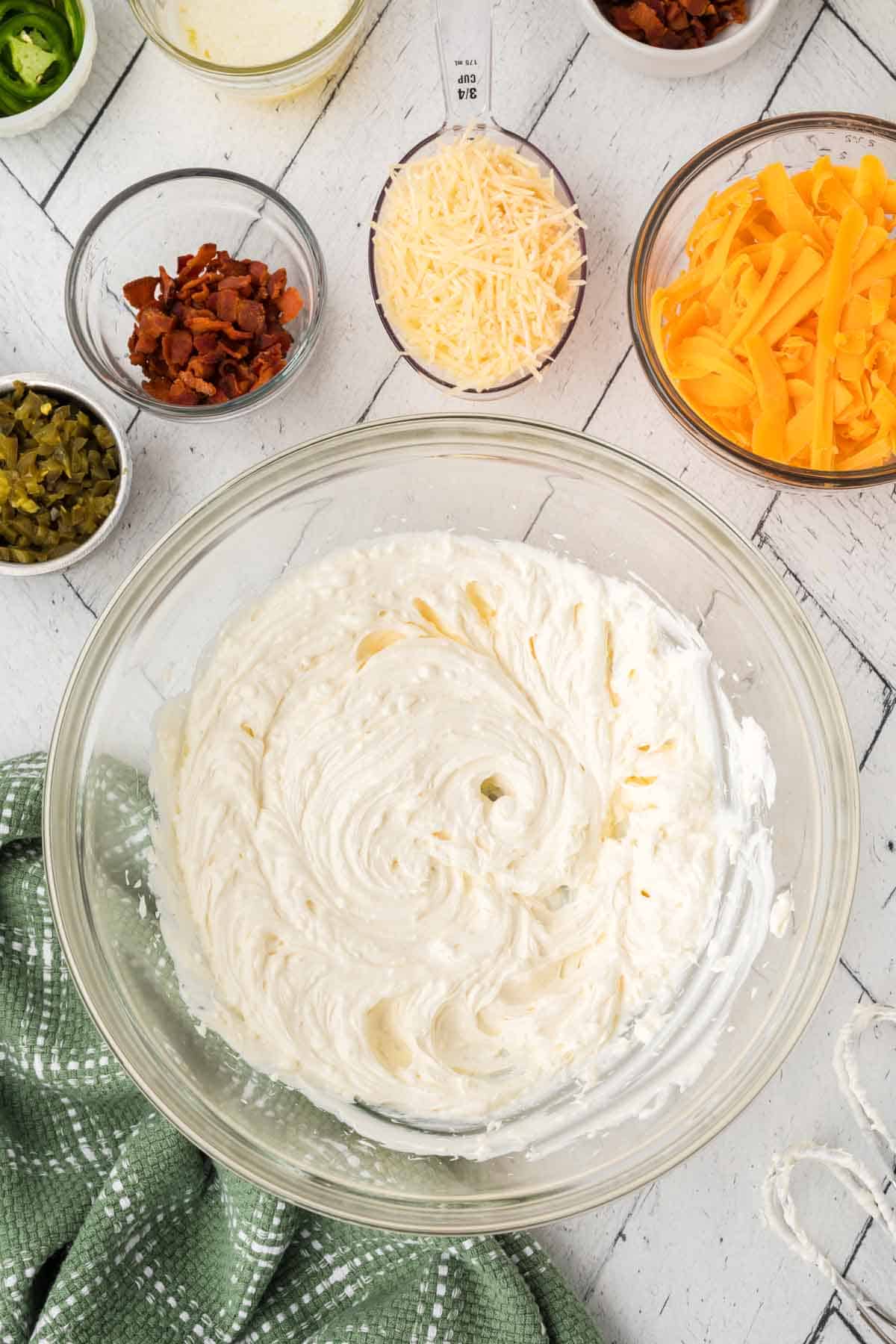 A large glass bowl filled with a creamy, white cheese mixture sits on a white wooden surface. Surrounding it are bowls of grated cheddar cheese, parmesan, chopped bacon, jalapeños, and sliced green onions—essential ingredients for the perfect Jalapeño Popper Dip Recipe. A green cloth is nearby.