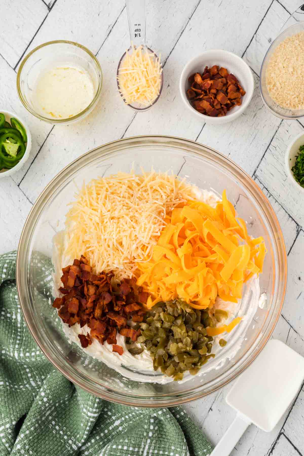 A glass bowl brimming with ingredients for a scrumptious jalapeño popper dip recipe: shredded cheese, chopped bacon, and diced jalapeños on a white tiled surface. Nearby are small bowls of additional toppings alongside a green cloth and spatula, ready to create magic.