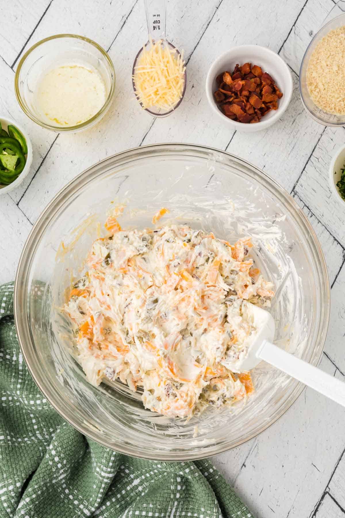 A glass bowl brimming with a creamy jalapeño popper dip recipe sits on a white wooden surface. Nearby, small bowls hold sauce, grated cheese, chopped nuts, and sliced jalapeños. A green checkered cloth peeks into view, promising a tasty adventure.