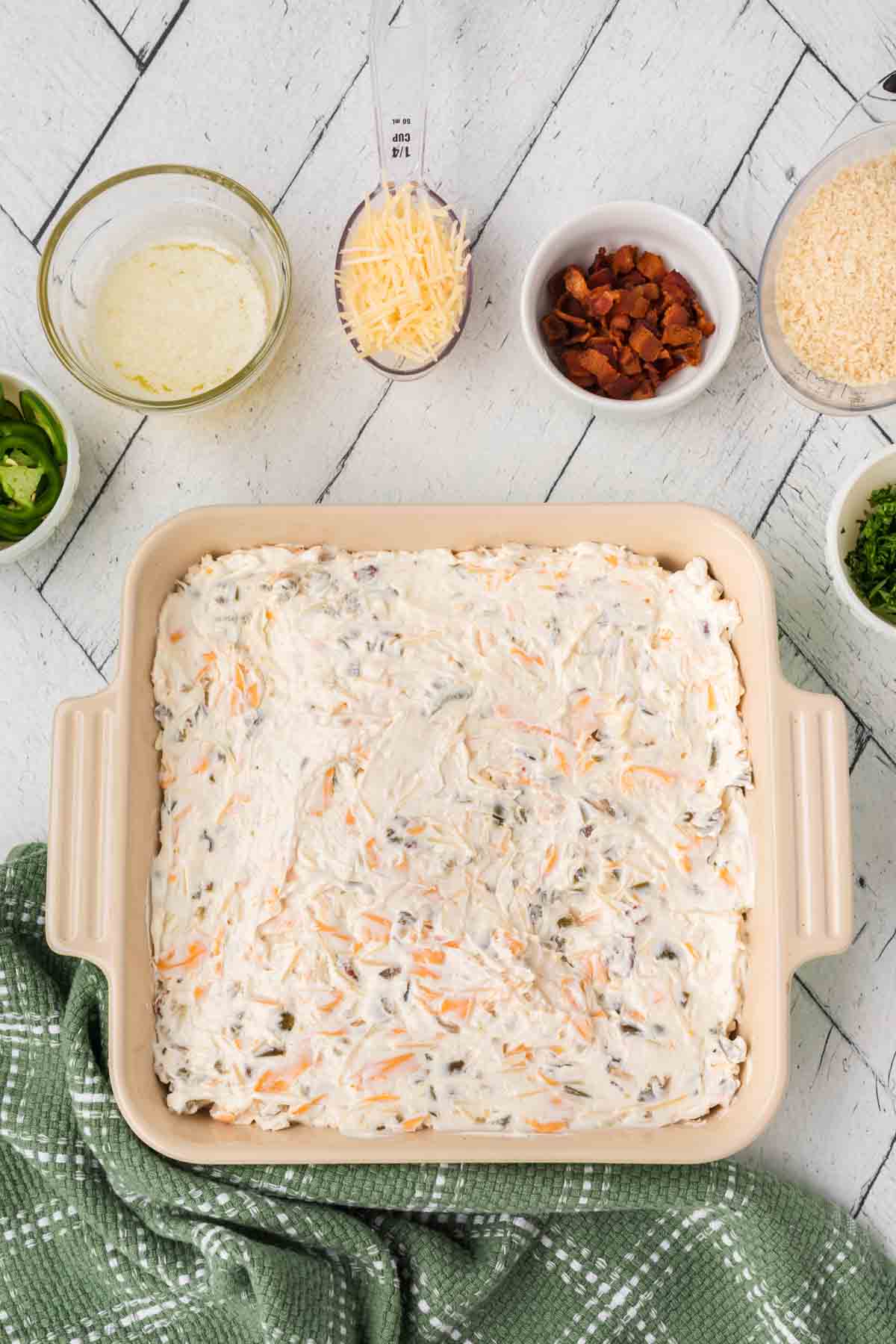 A square baking dish brimming with jalapeño popper dip is set on a white wooden surface. Nearby, small bowls and a measuring cup hold diverse ingredients like cheese, almonds, and herbs. A green kitchen towel adds a splash of color to this delectable scene.