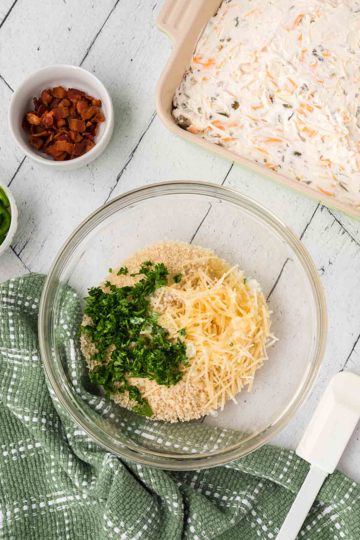 A glass bowl contains breadcrumbs, grated cheese, and parsley on a white wooden surface. Nearby is a dish with creamy Jalapeño Popper Dip, a small bowl of chopped bacon, and a spatula. A green kitchen towel is partially visible.