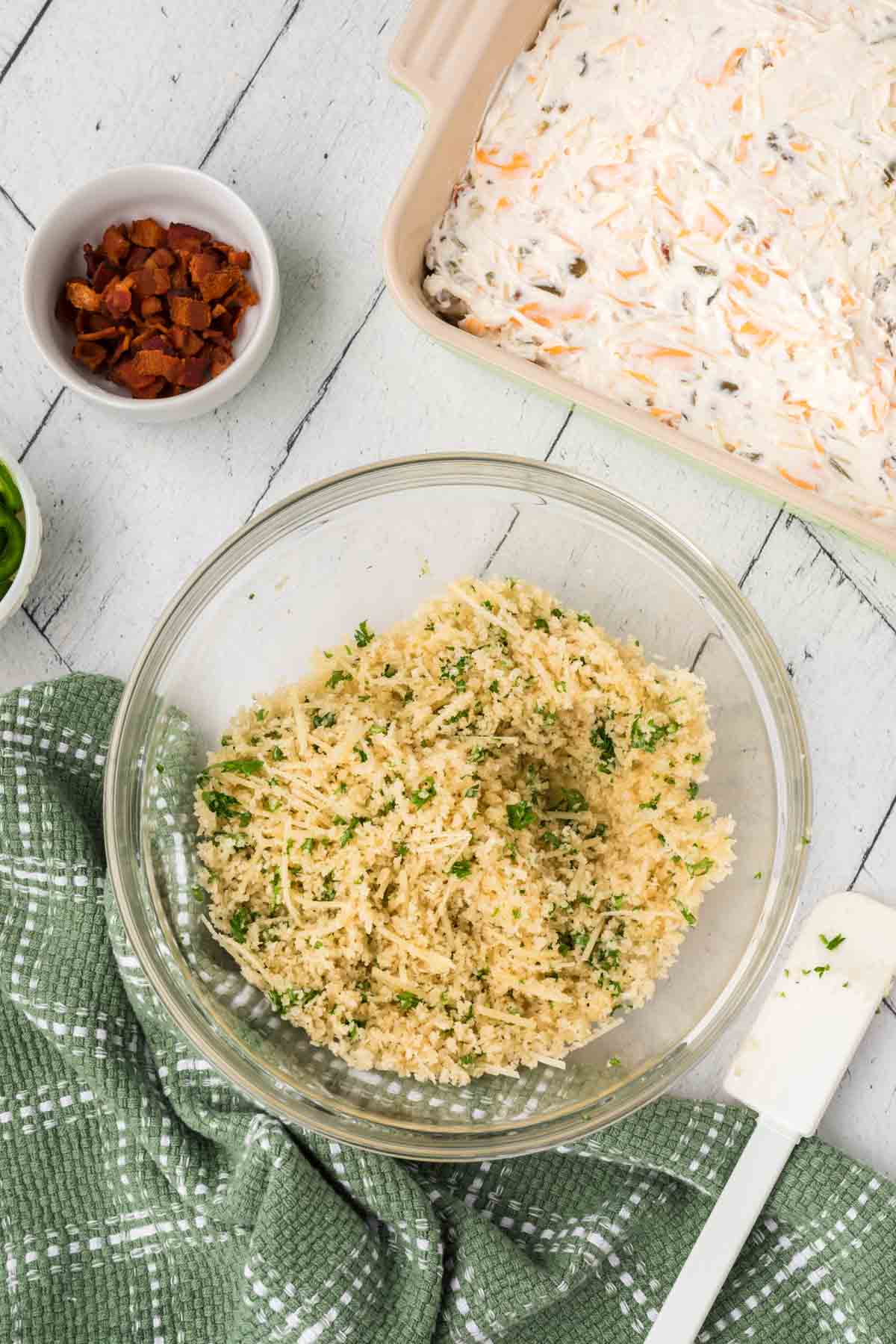 A bowl of couscous topped with chopped herbs sits on a white wooden surface. Nearby, a green kitchen towel accompanies a small bowl with diced bacon and an unbaked casserole dish filled with creamy jalapeño popper dip mixture. A white spatula rests beside the dish.