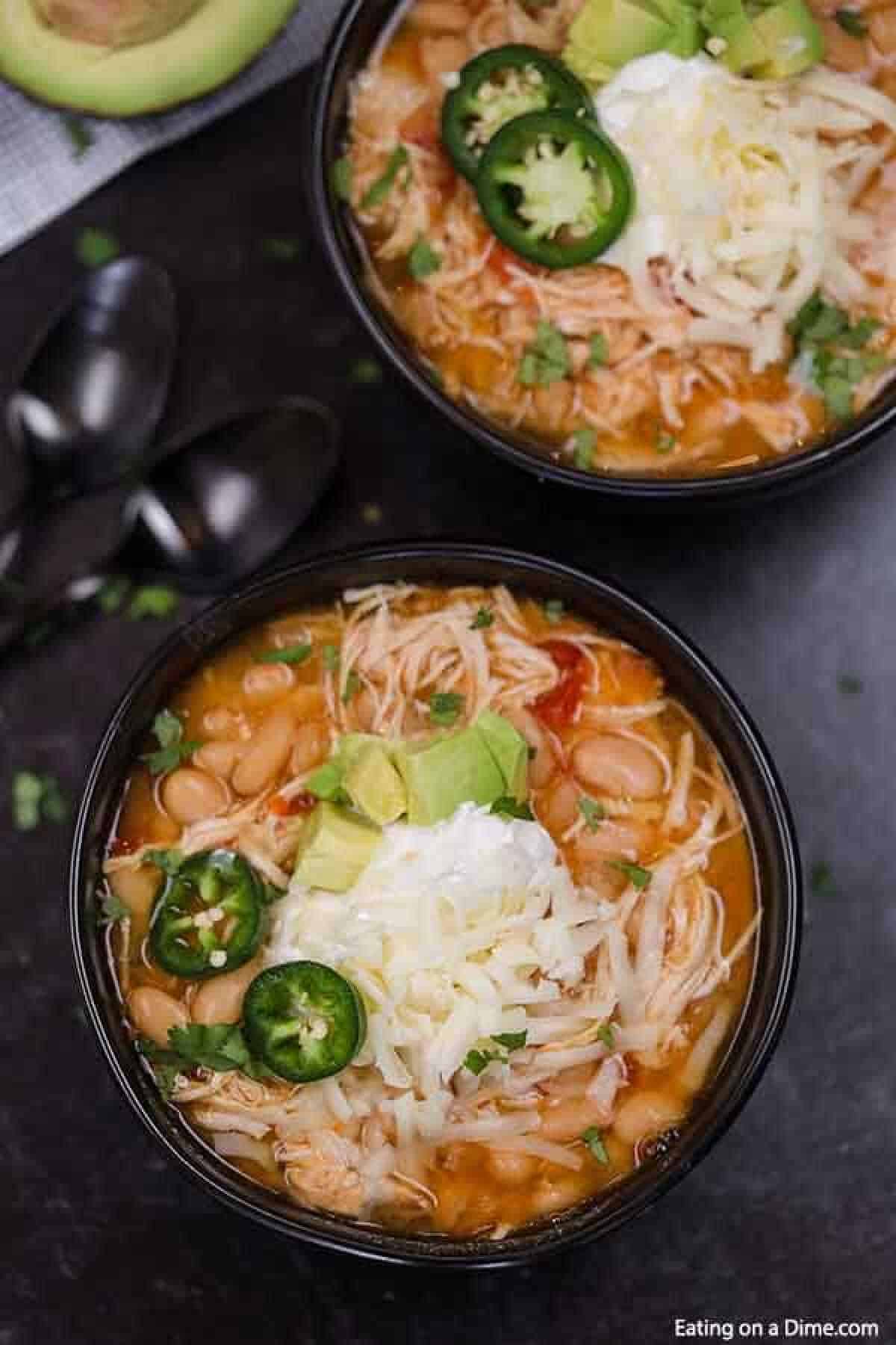 Two bowls of crock pot white chicken taco chili brimming with shredded chicken, white beans, and diced tomatoes are topped with cheese, jalapeños, sour cream, and avocado. Garnished with cilantro and paired with black spoons, this comforting dish is a flavorful feast.