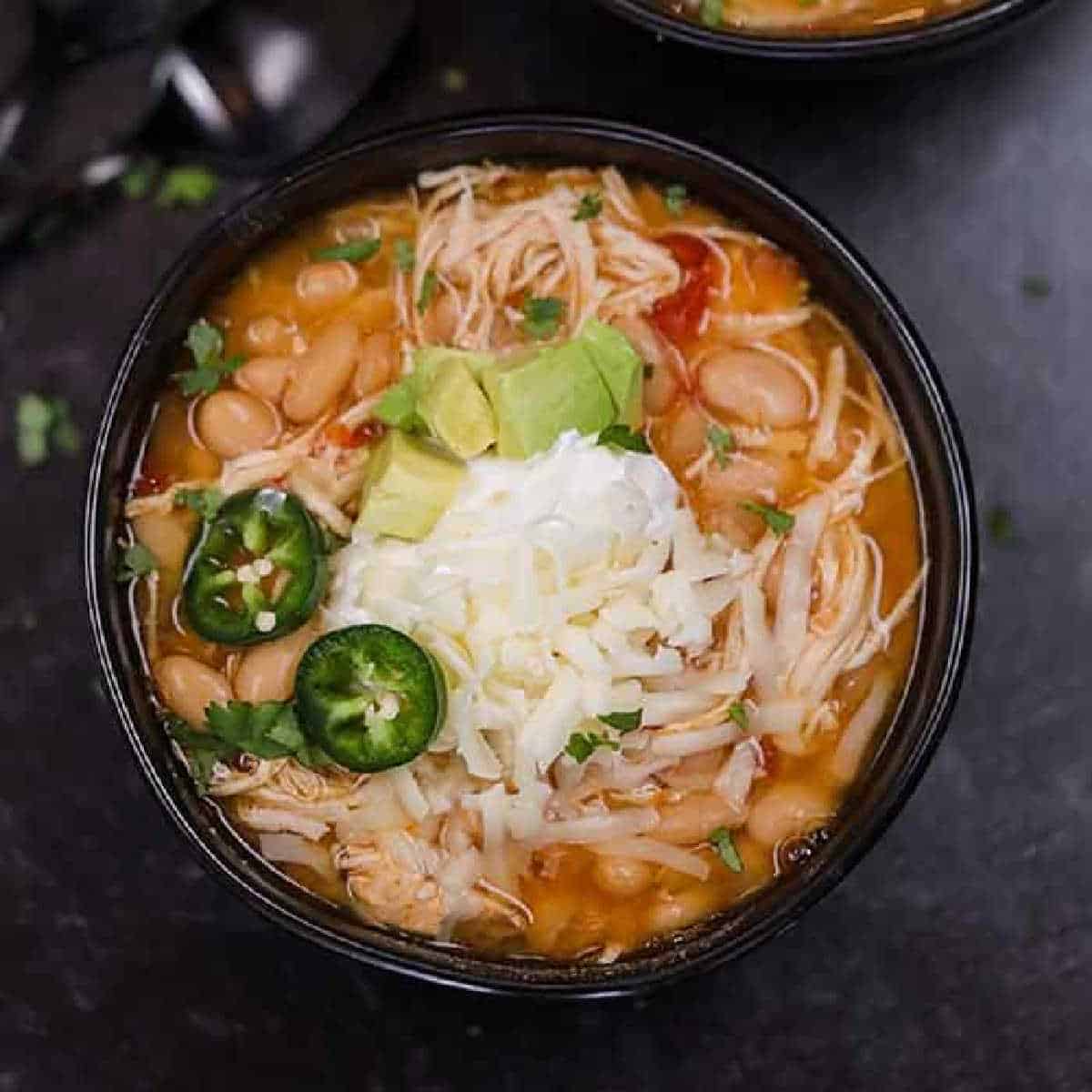 A crock pot white chicken taco chili features a bowl of soup brimming with shredded chicken, white beans, melted cheese, avocado slices, sour cream, and jalapeño slices. It's garnished with chopped cilantro. In the blurred background sit other bowls and spoons.