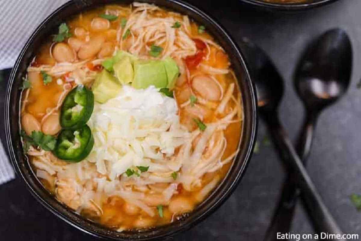 A bowl of crock pot white chicken chili topped with sliced jalapeños, shredded cheese, avocado, and a dollop of sour cream. Two black spoons are placed beside the tempting dish on a dark surface.