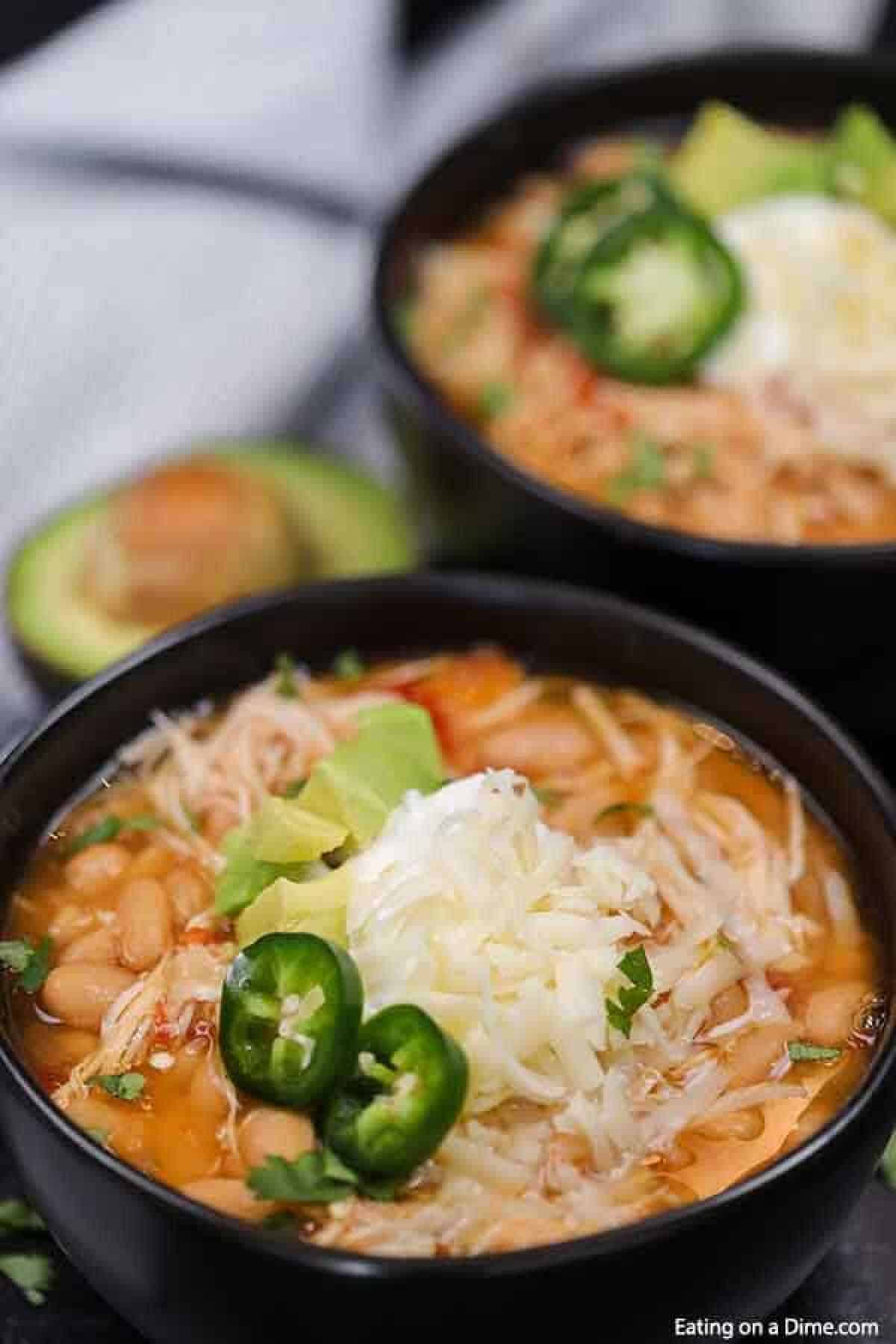 Crock pot white chicken chili soup topped with shredded cheese, sliced jalapeños, and avocado, garnished with cilantro, with a blurred second bowl and an avocado half in the background.