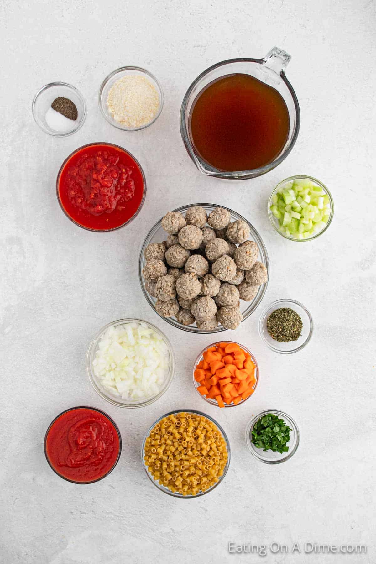 Top-down view of neatly arranged crock pot Italian meatball soup ingredients on a white surface. Includes raw meatballs, beef broth, diced tomatoes, tomato paste, celery, carrots, onions, pasta, parsley, seasoning, and spices in various bowls and measuring cups.