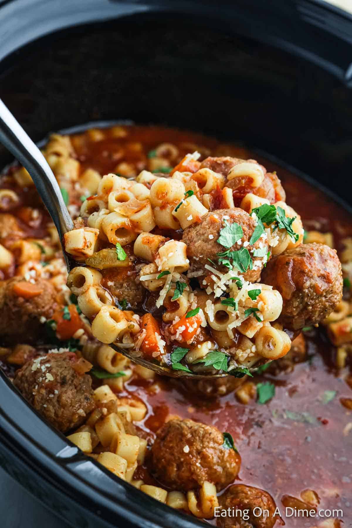 A ladle scoops Crock Pot Italian meatball soup, rich with pasta, meatballs, carrots, and tomatoes, garnished with grated cheese and fresh herbs.