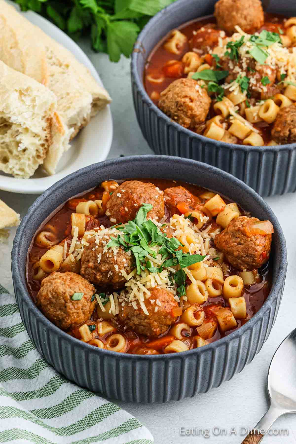 Two gray bowls brim with pasta, meatballs, and a rich red sauce reminiscent of crock pot Italian meatball soup, topped with grated cheese and fresh herbs. Slices of bread rest on a nearby plate, accompanied by a striped napkin and vibrant green parsley in the background.