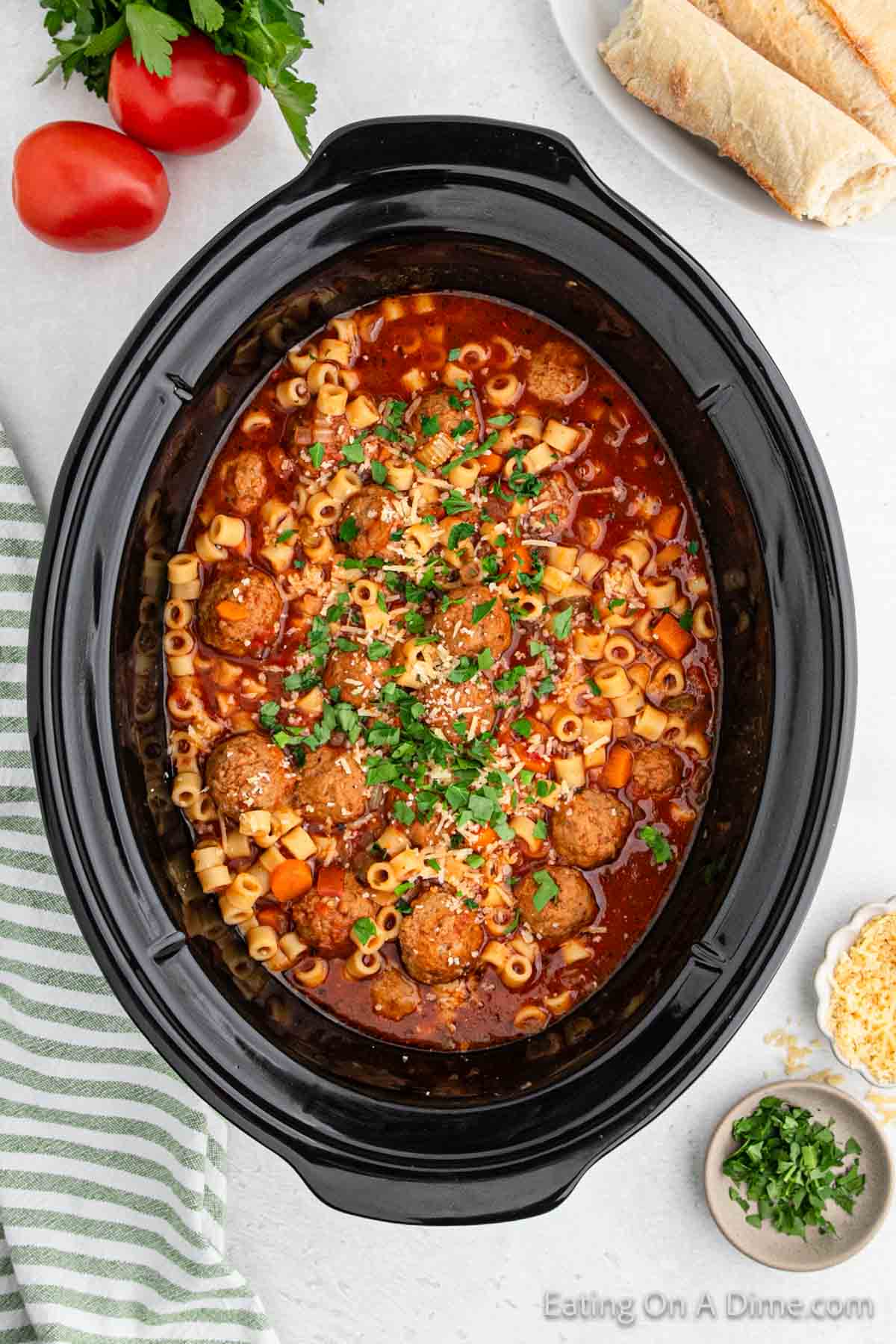 A crock pot brimming with Italian meatball soup showcases pasta, juicy meatballs, and carrots in a rich tomato broth, topped with chopped parsley and shredded cheese. Nearby sit fresh tomatoes, parsley, cheese, and bread.