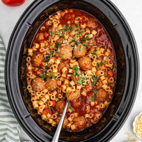 A crock pot filled with pasta, Italian meatball soup, and a rich tomato sauce. The dish is garnished with chopped parsley and grated cheese. A metal serving spoon is placed inside, ready to serve.