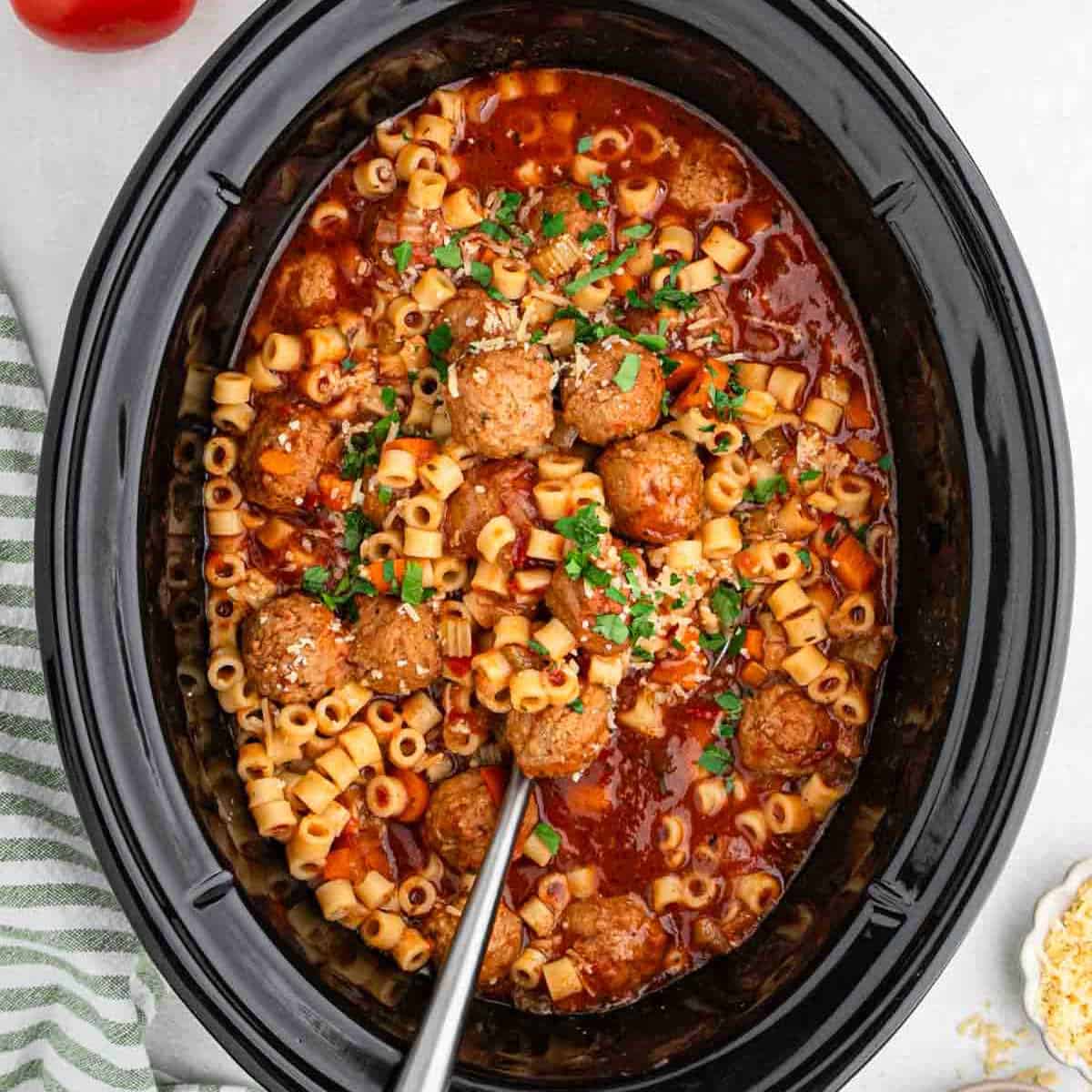 A crock pot filled with pasta, Italian meatball soup, and a rich tomato sauce. The dish is garnished with chopped parsley and grated cheese. A metal serving spoon is placed inside, ready to serve.