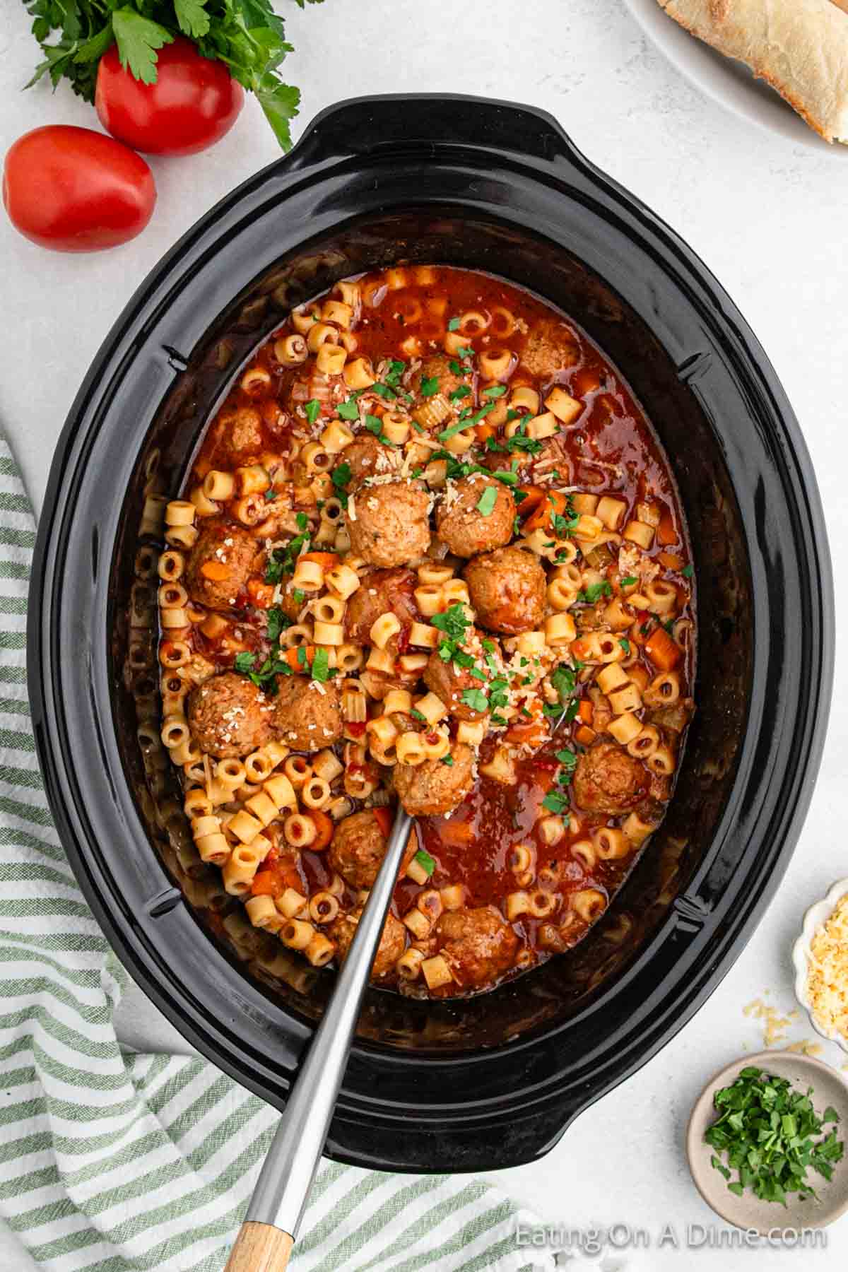 A crockpot brimming with Italian meatball soup, featuring pasta and tomato sauce, garnished with parsley. A ladle rests inside. Surrounding are fresh parsley, ripe tomatoes, grated cheese, and a striped napkin on a light-colored table.