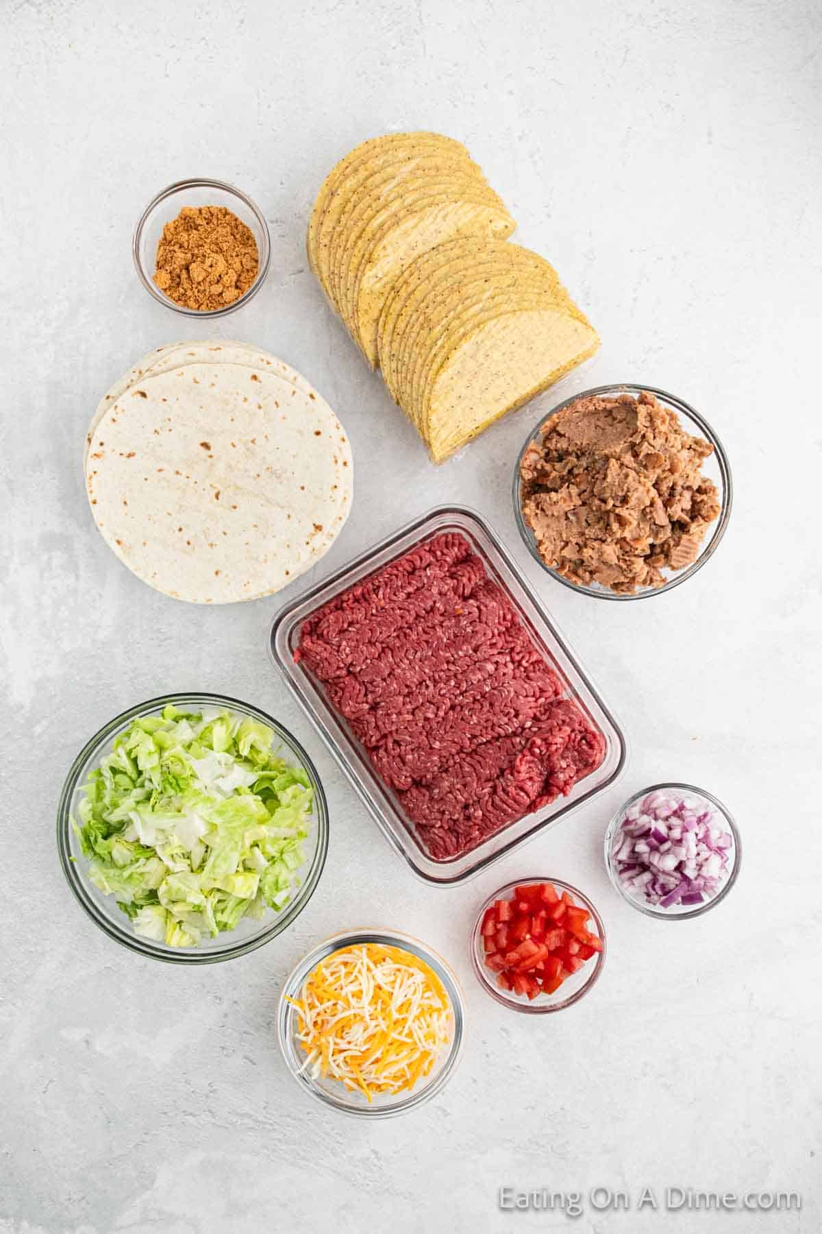 Ingredients for a Double Decker Taco Recipe are arranged on a white surface, featuring ground beef, refried beans, taco seasoning, hard and soft taco shells, shredded cheese, lettuce, diced tomatoes, and chopped onions.