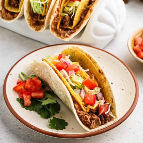 A Double Decker Taco on a plate, bursting with ground meat, shredded cheese, diced tomatoes, and lettuce. Fresh chopped tomatoes and cilantro garnish the vibrant dish. Other tacos are artfully arranged in the background on a holder, showcasing this delicious recipe.