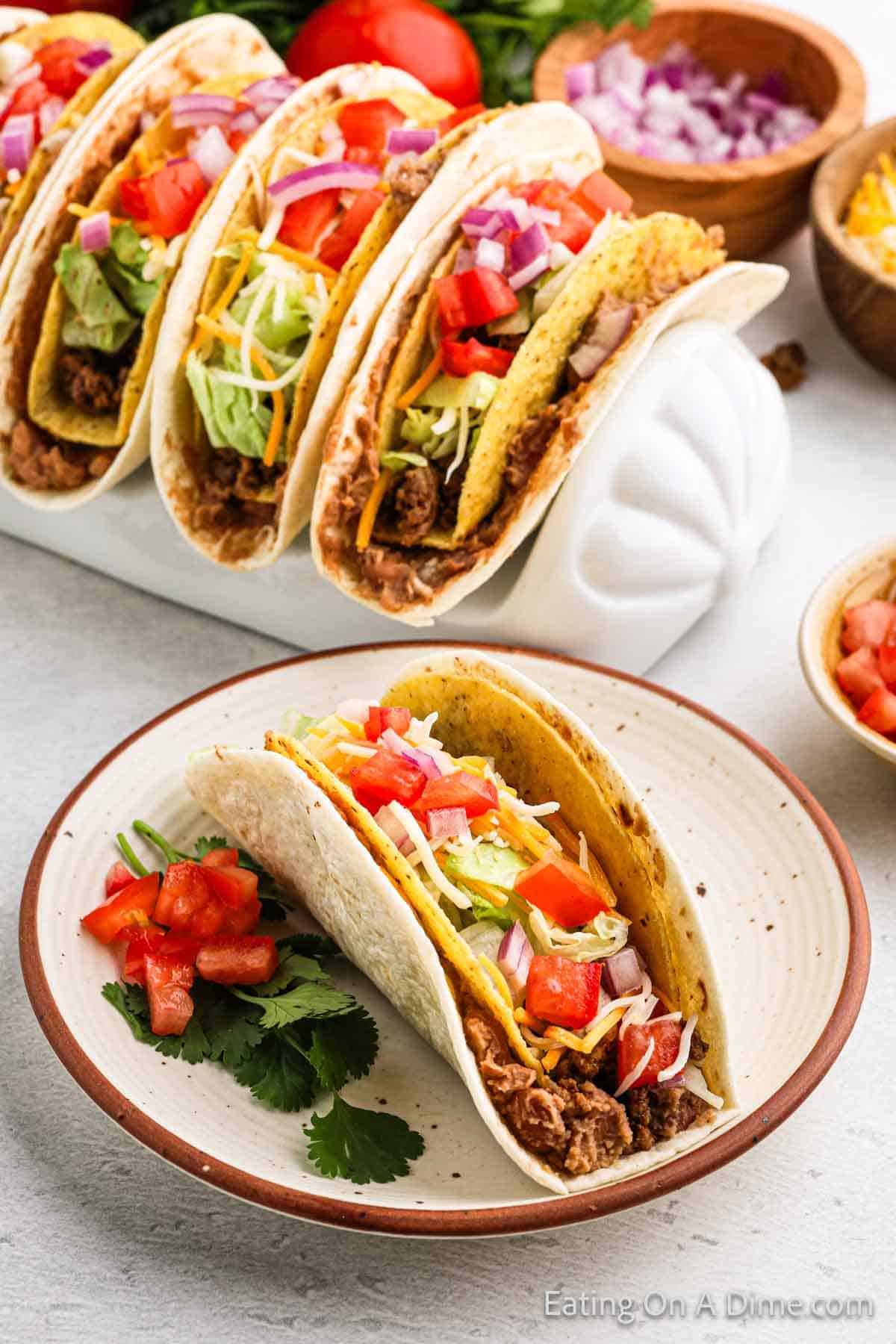 A plate with two tacos filled with ground beef, lettuce, cheese, tomatoes, and onions sits temptingly. In the background, more tacos rest in a taco holder alongside fresh ingredients like chopped onion and cilantro—perfect for creating your own Double Decker Taco recipe masterpiece.