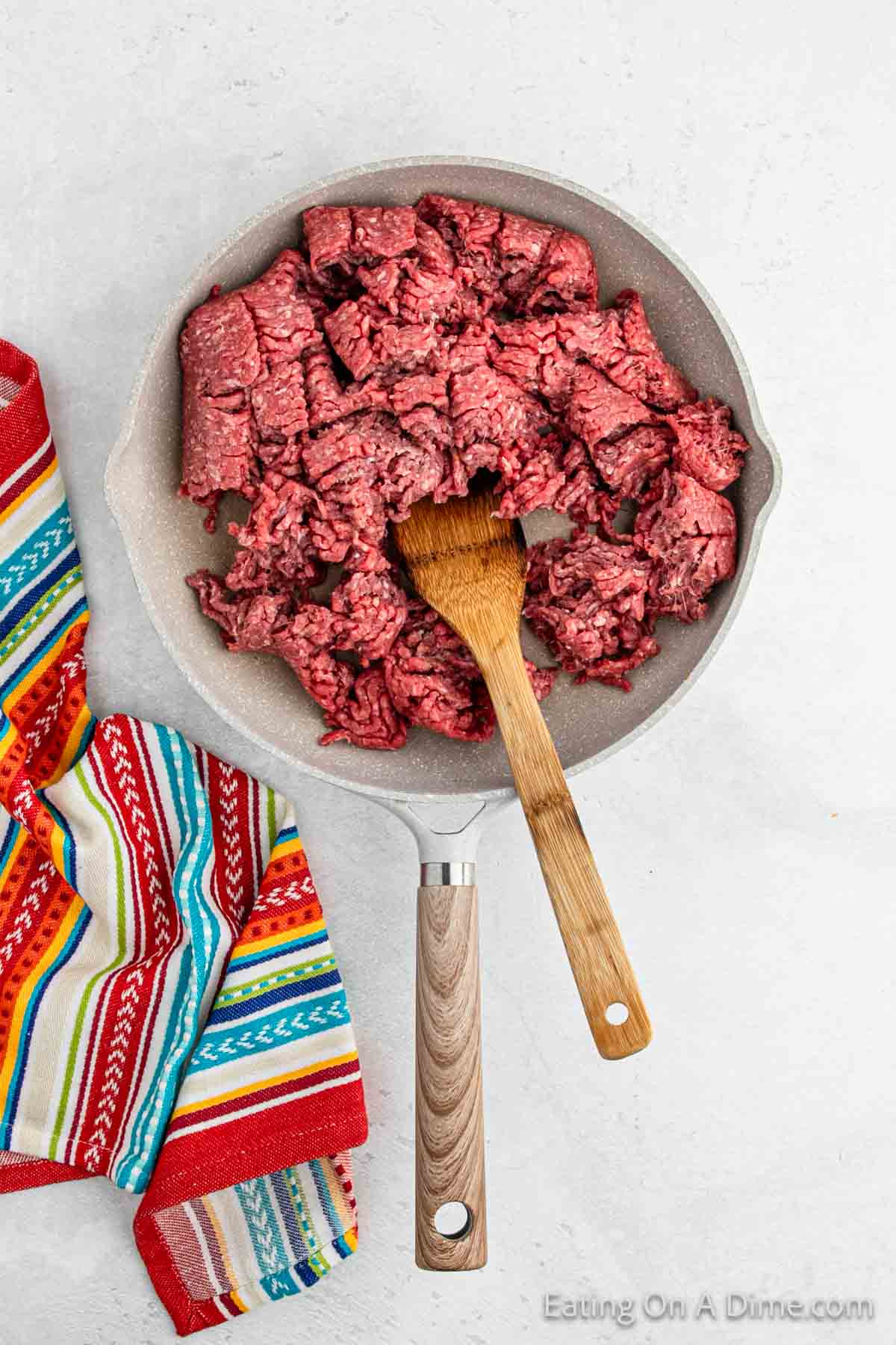 A frying pan contains raw ground beef, ready to be transformed into a delicious Double Decker Taco recipe, with a wooden spatula resting inside. Next to the pan, there's a colorful, striped cloth on a light gray surface.