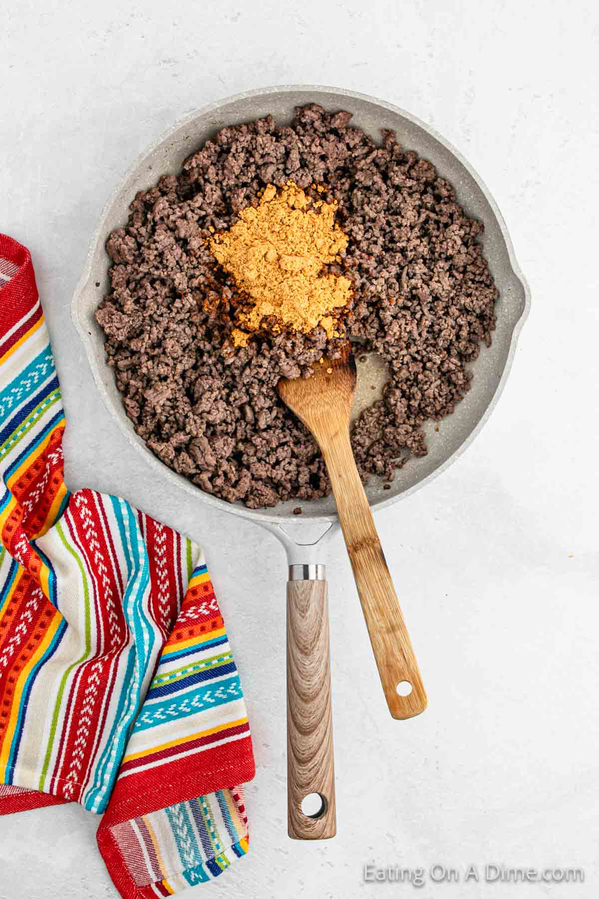 A frying pan with browned ground beef and taco seasoning in the center, stirred by a wooden spoon, perfect for a double-decker taco recipe. A colorful striped fabric is placed on the side.