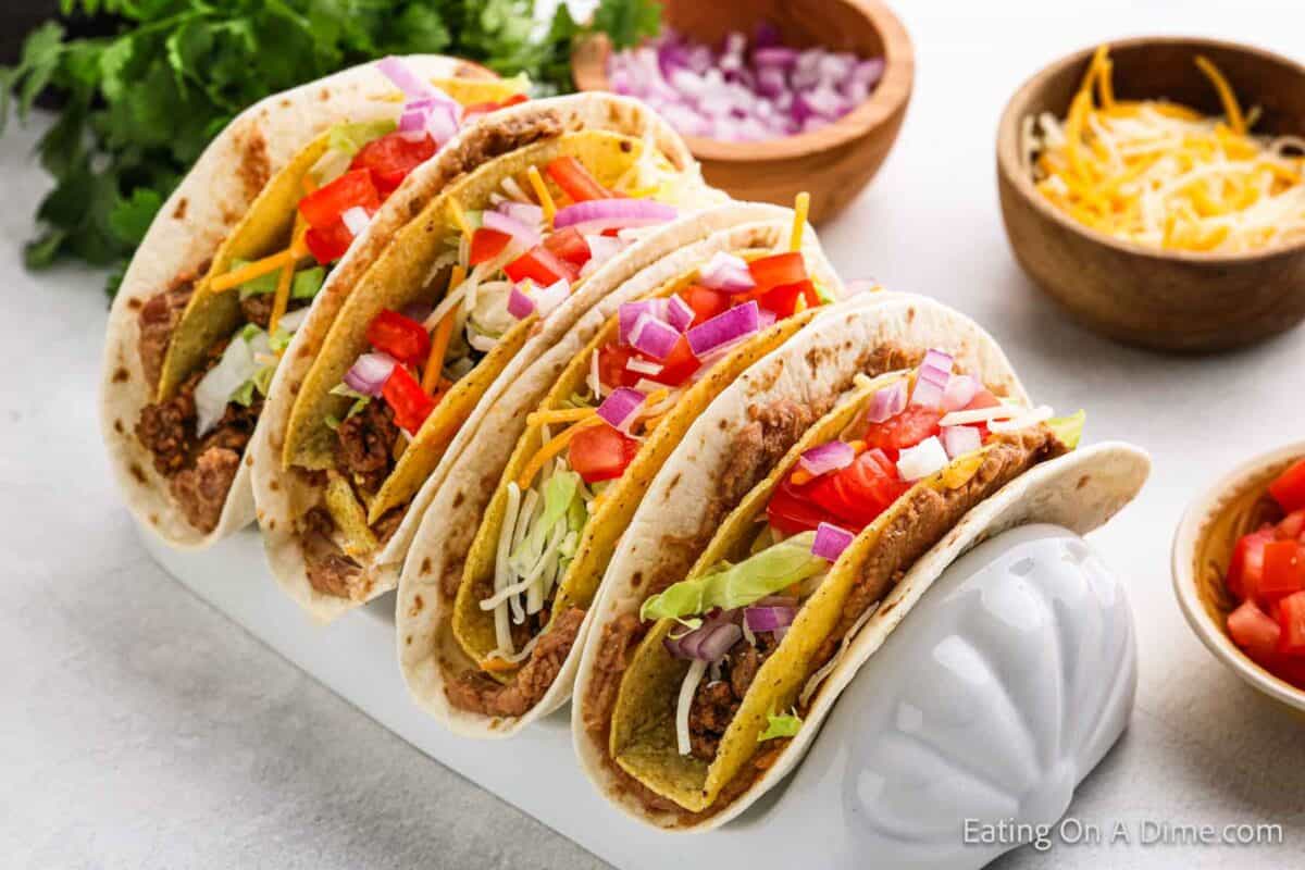 Four tacos filled with seasoned meat, shredded cheese, lettuce, diced tomatoes, and onions are neatly arranged in a taco holder. Inspired by a double decker taco recipe, bowls of diced onions, shredded cheese, and fresh cilantro are visible in the background.